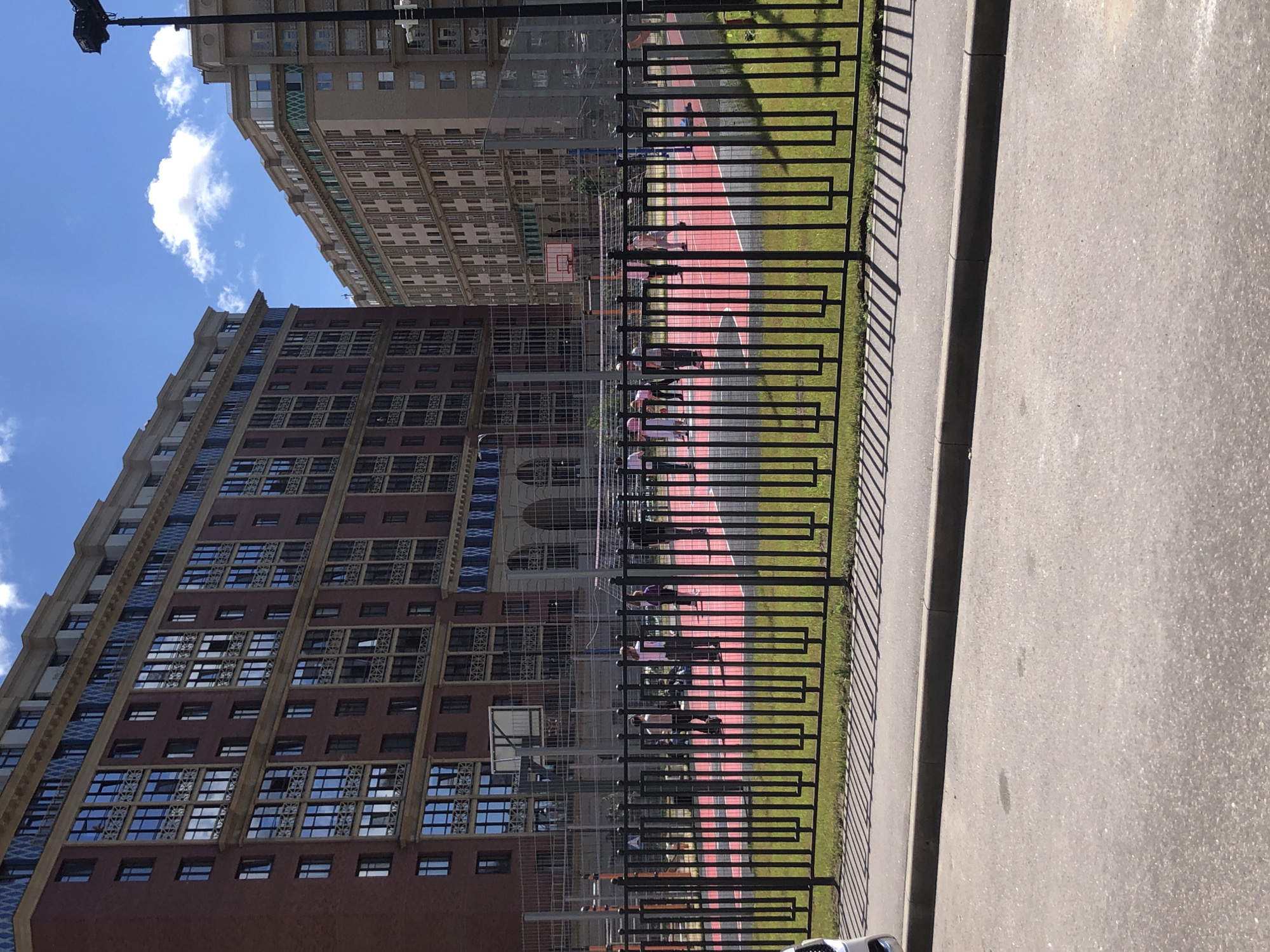 This image captures an outdoor scene in Saint Petersburg, Russia, under a bright, sunny sky with scattered clouds. The perspective is tilted, giving a dynamic view of urban architecture and recreational space.
In the foreground, a paved pathway runs alongside a black metal fence, casting distinct shadows from the sunlight. Beyond the fence is a bright red sports court, possibly a basketball or tennis court, with vibrant green grass bordering it. Several individuals, dressed in casual attire, are visible on the court, engaging in what appears to be recreational activity or practice. Some are in motion, suggesting a game or training session is in progress. The figures are silhouetted against the bright court, making detailed identification difficult.
Towering above the court and fence are two large, multi-story buildings. The building closer to the foreground is a dark red brick structure with numerous windows, many reflecting the blue sky. Its architecture features decorative elements between the rows of windows. The building behind it is lighter in color, possibly beige or light brown, and also exhibits a patterned facade with many windows. The scale of these buildings suggests a dense urban environment, characteristic of a major city like Saint Petersburg.
The overall impression is one of a bright, active day in an urban setting, where modern architecture coexists with outdoor recreational facilities. The clear sky and strong sunlight indicate it is daytime, likely midday, given the length and direction of the shadows. There is no visible text in the image.