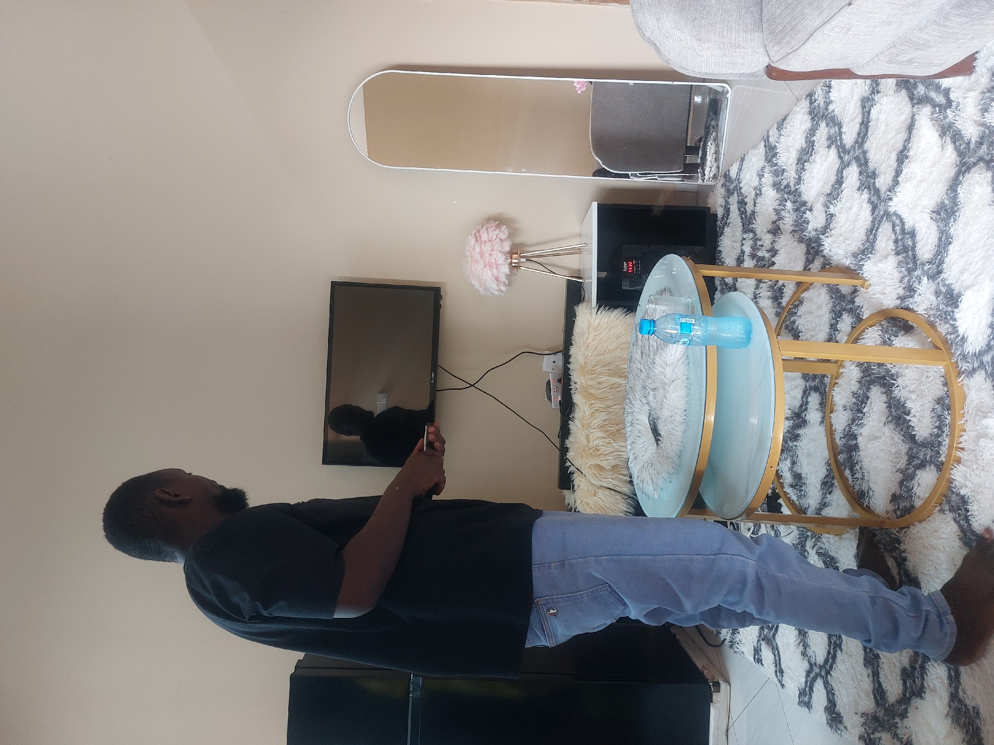 A man wearing a black t-shirt and light blue jeans stands barefoot in a room. He is holding a mobile phone. Behind him, a black television is mounted on the wall. Above the television is a white-framed mirror, and to the side is a white table with a lamp. A two-tiered glass table with gold legs stands near the television and is partially covered by a white fur throw. Two bottles of water are on the table. The room is carpeted with a white rug with a black and gray geometric pattern. The location appears to be a residential interior. There is no indication of the city of Dar es-Salaam, Tanzania, or any specific location.