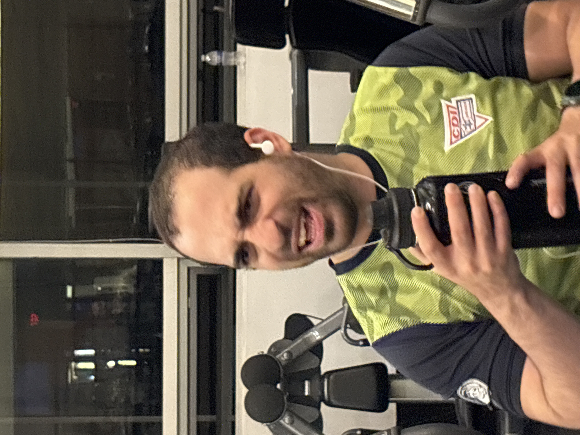 A man with dark hair and a light beard is holding a black water bottle and looking at the camera with a strained expression. He is wearing a light green camouflage shirt with a "CDII" logo on the upper right chest and a blue stripe that separates the camo from the black sleeves, with what appears to be a logo on the lower part of the sleeves. He also has a white wireless earbud in his right ear and is wearing a wristwatch on his left wrist. The background includes a window showing a city scene at night with some lights and buildings, and a piece of gym equipment. It appears to be in Mexico City based on the CDI logo on the shirt.