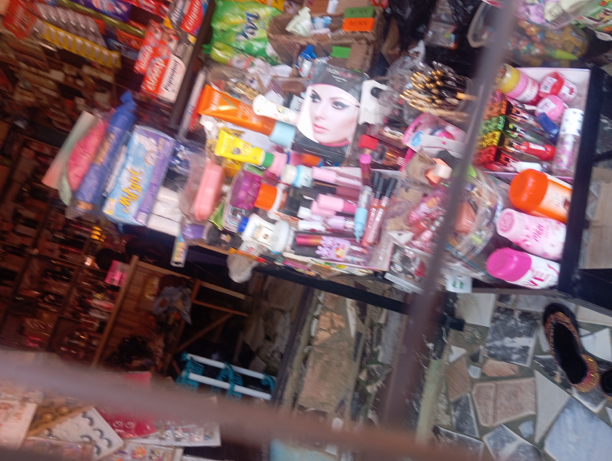 The image depicts a vendor's stall in Nkpor, Nigeria, displaying a variety of cosmetic and personal care products. Items visible include tubes of toothpaste, packaged sweets, tubes of creams and lotions, lipsticks, eyeliners, and other makeup items. The stall is stocked with a wide assortment of goods, typical of street-side commerce in the area. The environment appears cluttered. A sandal rests on the ground beside the display. The stall setup, with its assortment of small items, suggests a common commercial scene in Nkpor.