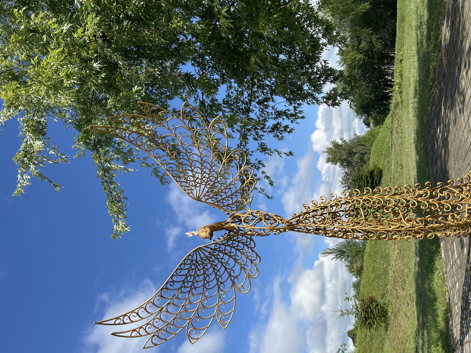 The image depicts a golden, openwork sculpture of a bird, possibly a phoenix, standing in a park-like setting. The sculpture is intricately detailed with swirling patterns forming its body and wings. It is positioned on a paved area, likely a pathway, with a grassy hill and trees in the background. The sky is a vibrant blue with scattered white clouds. To the right of the sculpture, a leafy green tree branches overhead. The overall impression is one of a bright, sunny day in an outdoor public space. The sculpture is the primary subject, a decorative element within a natural landscape. There are no people or animals visible in the image. The location is identified as Азярэцкі сельскі Савет, Belarus.
