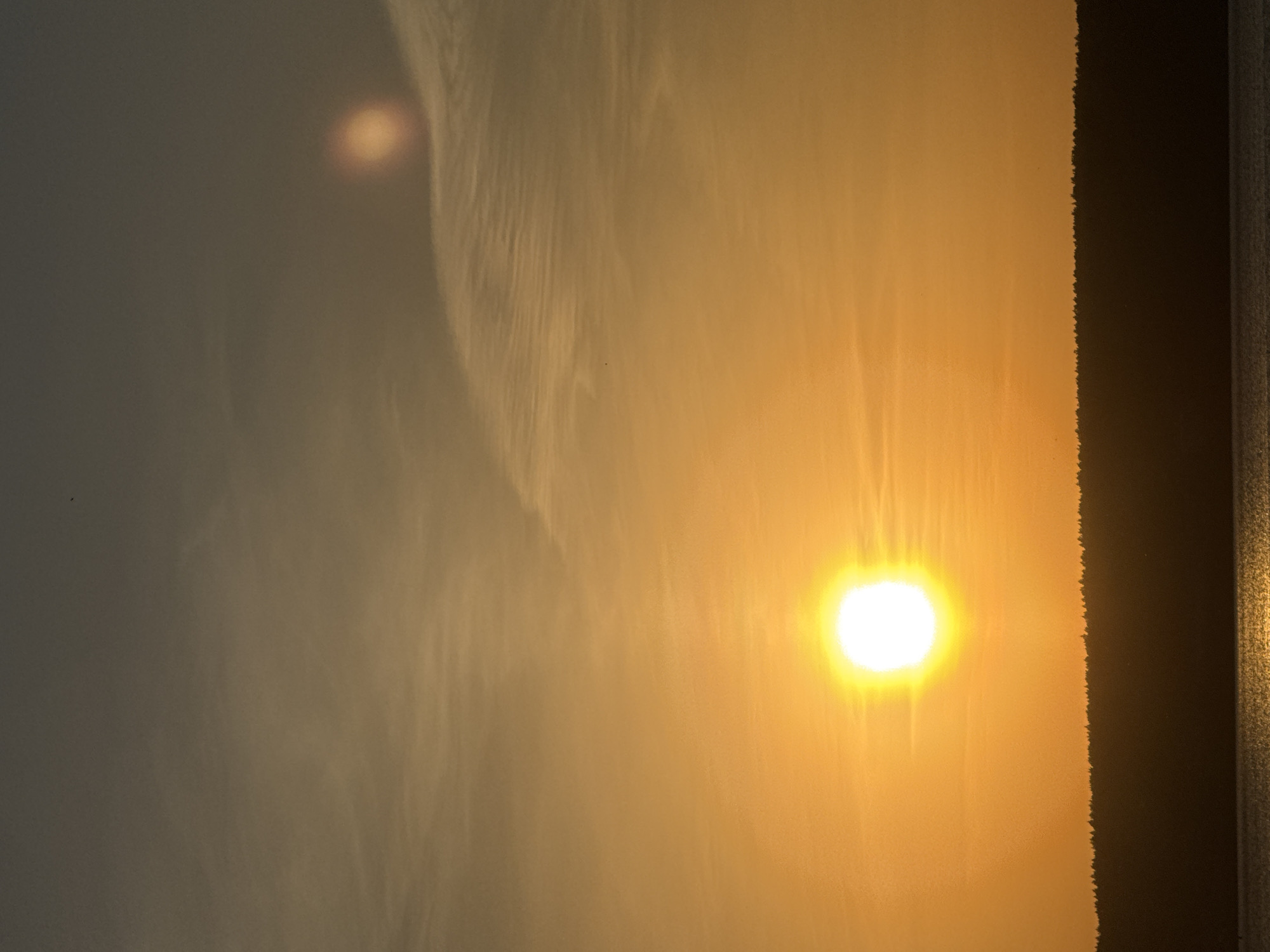 The image shows a sun setting or rising through a window. The light is a bright, diffused yellow-orange, and the sky appears to be hazy or filled with clouds. A dark frame of the window is visible on the right side of the image. There is no indication in the image it was taken in Naberezhnye Chelny, Russia, or any other specific location. The image includes no people, subjects, objects, actions or interactions that can be identified.