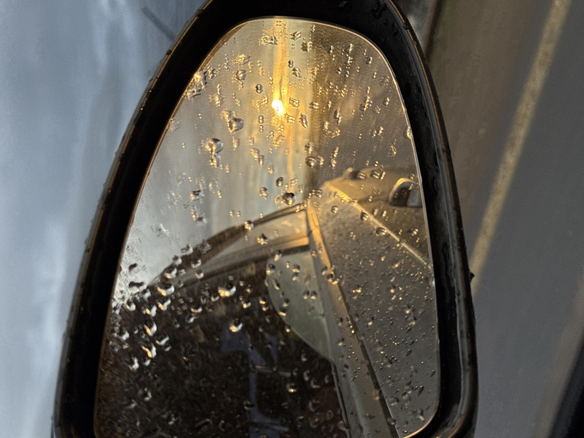 A close-up image captures a car's side-view mirror, prominently covered with numerous water droplets, indicating recent rain or high humidity. The mirror reflects a radiant, golden light, suggesting either sunrise or sunset, which creates a warm, intense vertical glow against the otherwise muted tones. Parts of the car's body, including a section of its door and frame, are visible in the reflection at the bottom and right of the mirror. To the right of the mirror's frame, a grey surface resembling a road or paved area, possibly with a faint line, can be seen. The overall setting is outdoors, likely from inside a parked or moving vehicle. There is no visible text in the image. This scene was captured in Chaikovsky, Russia.