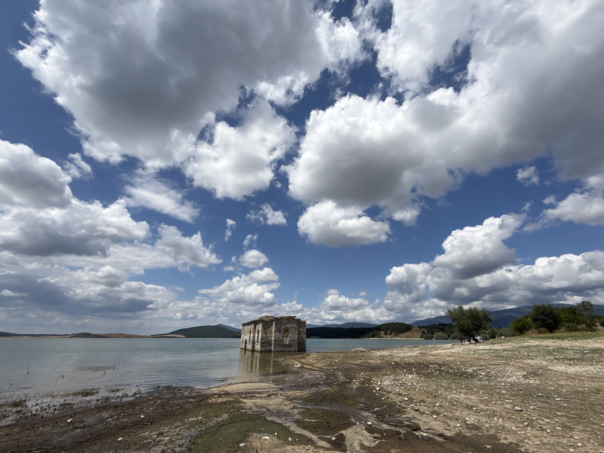 A wide-angle shot captures the partially submerged St. John the Baptist Church, a stone structure rising from the tranquil waters of Lake Zhrebchevo in Panicherevo, Bulgaria. The sky is a dynamic expanse of blue, dotted with dramatic, fluffy white and grey cumulus clouds. The lake's surface is calm, reflecting the sky and surrounding landscape. The foreground is a muddy, dry shoreline littered with pebbles, suggesting a receding water level. In the mid-ground, the abandoned church stands as the central focus, its stone walls weathered and its roof missing, with windows showing a darker interior. To the right of the church, a small cluster of trees and vegetation borders the lake. In the distance, rolling hills covered in green foliage and darker green forested mountains form the horizon. The overall scene depicts a serene yet somewhat desolate landscape, highlighting the contrast between the ancient architecture and the vast natural environment.