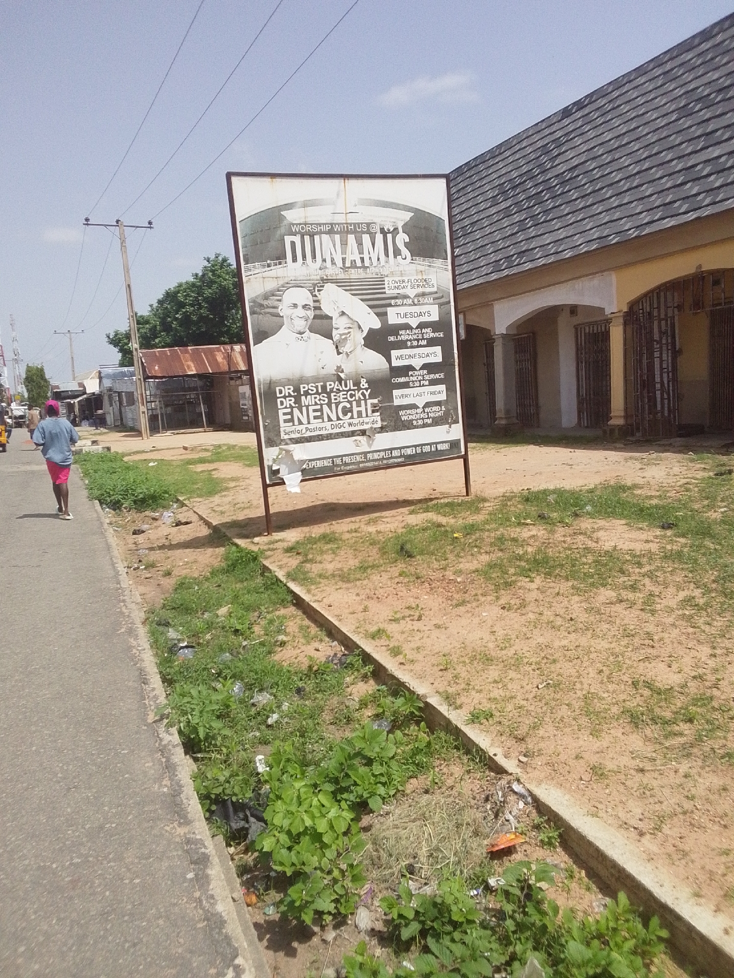 The image depicts a street scene in Jalingo, Nigeria, under a clear, bright sky, suggesting it is daytime. A large billboard advertising a church, "Dunamis," is prominently displayed on the right side of the road. The billboard features images of two individuals, Dr. Pst Paul and Dr. Mrs Becky Enenche, identified as Senior Pastors of DIGC Worldwide. It also lists various service times, including Sunday services, Tuesday healing and deliverance services, Wednesday power communion services, and a "Worship, Word & Wonders Night" held on the last Friday of the month.
To the left of the billboard, a person wearing a blue t-shirt and pink shorts walks away from the camera along a paved road. Further down the road, several buildings with corrugated iron roofs and metal gates are visible, indicating a commercial or residential area. The foreground of the scene is a patch of dry, sandy ground with sparse green vegetation, and some litter is scattered along the edge of the road and in the grassy area. Overhead, power lines are strung between utility poles, a common sight in many urban and semi-urban environments. The overall impression is a typical street view in a Nigerian town, with a mix of religious, commercial, and everyday elements.