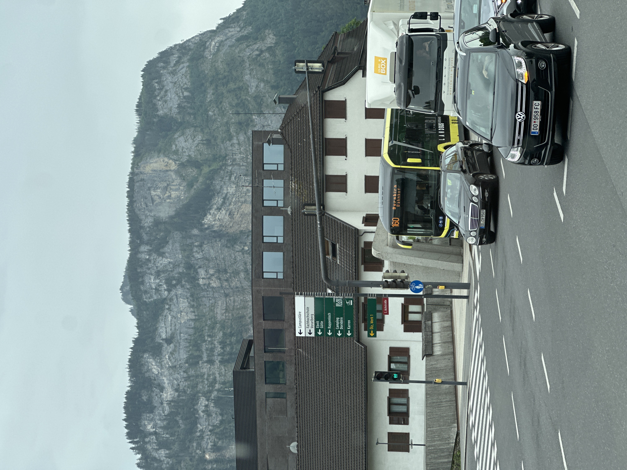 The image shows a street scene in Dornbirn, Austria. A traffic intersection is visible with cars, a bus, and a truck. A signpost in the center-left of the image indicates directions to various locations within Dornbirn, including "CampusVärë," "Fachhochschule Vorarlberg," "Ebnit Gütle," "Rappenloch," "Camping Dornbirn," "Karren," and "Ötz. Werk 1." A traffic light with a green pedestrian signal is also visible. In the background, a large, rocky mountain with trees covers the skyline. The sky is overcast.