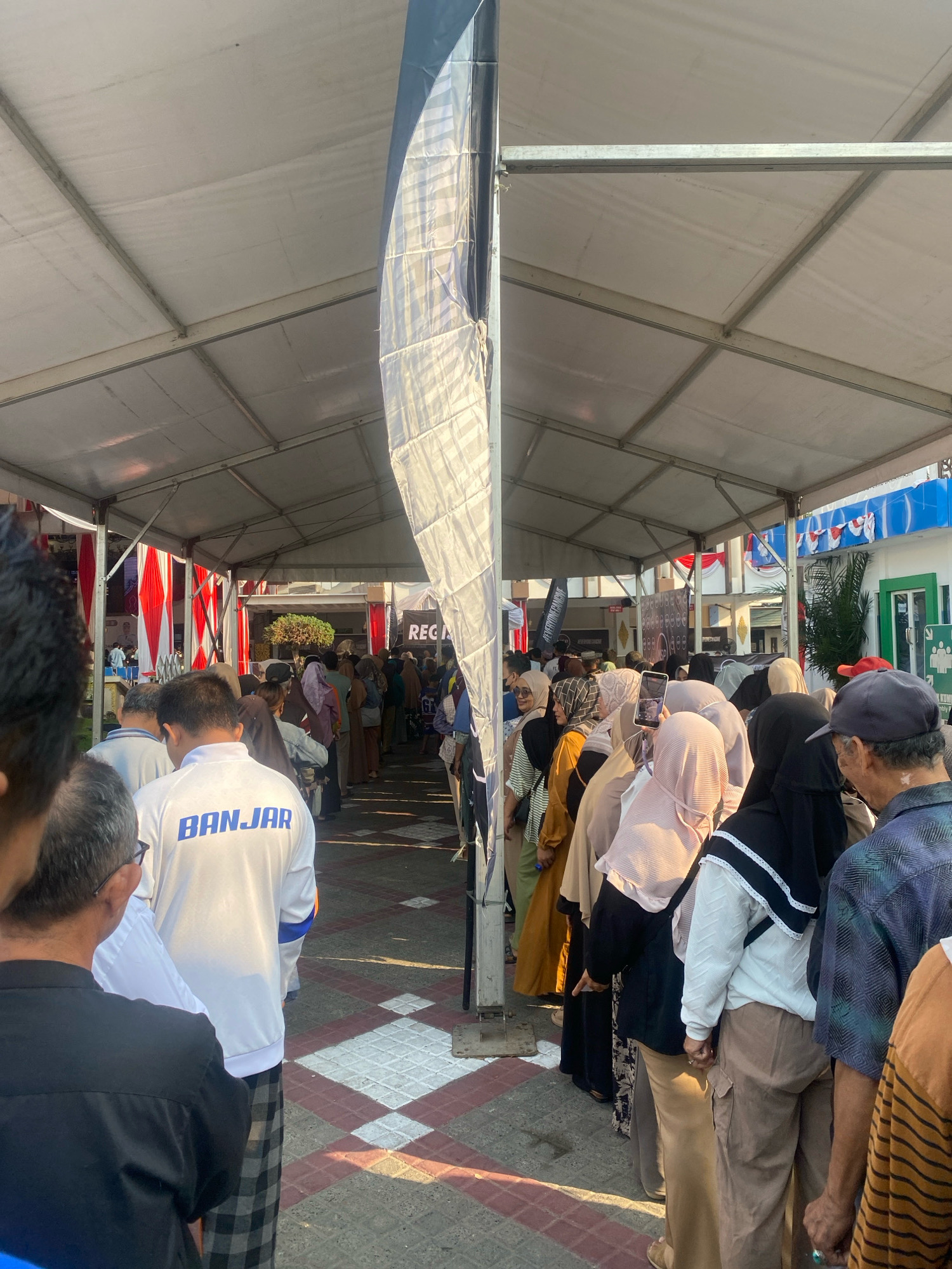 The image captures a scene outdoors, possibly in the city of Banjarmasin, Indonesia, under a large white tent structure. A long queue of people, predominantly women wearing hijabs and men in casual attire, are lining up. Some individuals are holding phones, and one woman is taking a photograph. The flooring features a pattern of white and reddish-brown diamond shapes. Red and white striped banners are visible on the left, and a large banner with the word "REG" partially visible is in the background. The sunlight casts shadows, suggesting it is daytime. One person in the foreground is wearing a white jacket with "BANJAR" printed in blue on the back. The overall atmosphere appears to be one of order and waiting, possibly for an event or service.