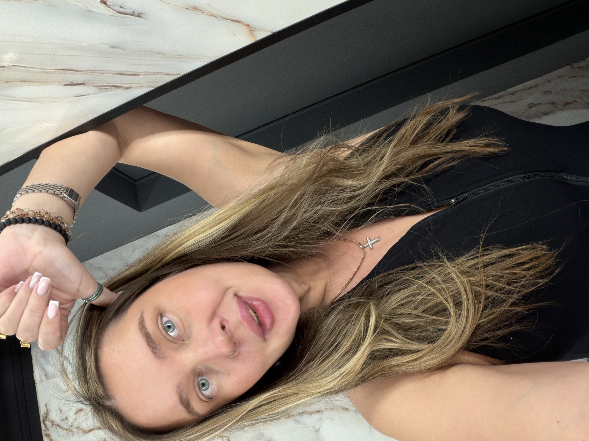A woman with light hair and blue eyes is lying down, possibly on a white marble surface with black accents. She is wearing a black top with a zipper. She has a cross necklace and several bracelets on her left wrist. Her left hand is near her face, with a green ring on one finger and a gold ring on another. The woman is looking at the camera and smiling. The image does not provide any evidence to identify the specific location within Moscow, Russia.