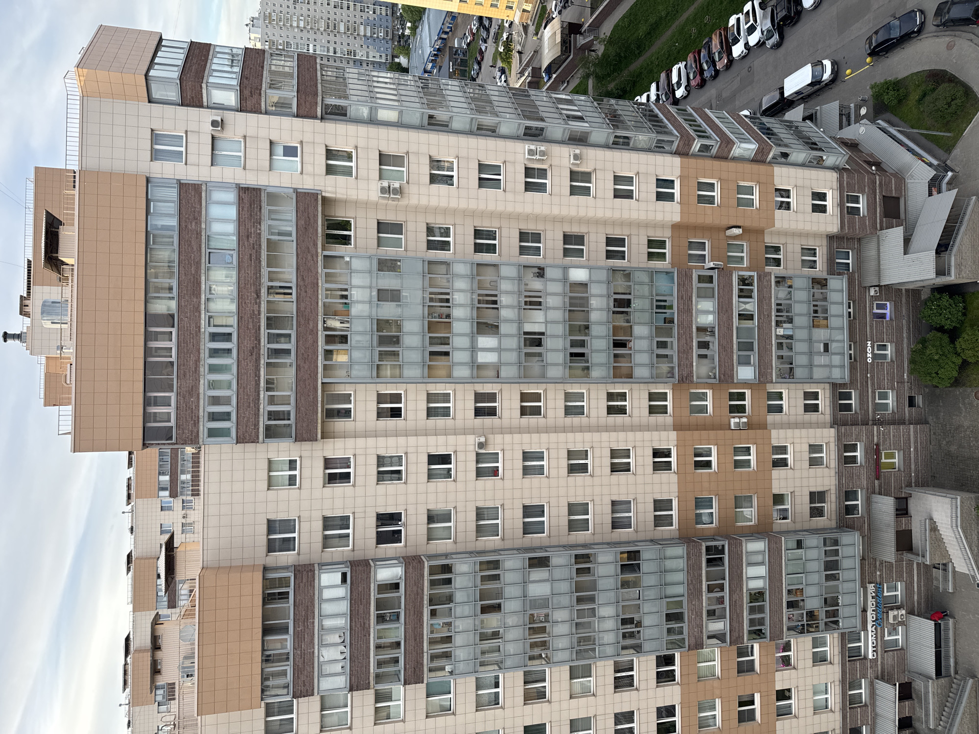This elevated view captures a modern, multi-story building under an overcast daytime sky. The building's facade is a mosaic of light beige and brown rectangular panels, complemented by sections featuring a reddish-brown brick-like texture. It boasts numerous windows of varying sizes, along with extensive glass-enclosed balconies or architectural features, including some sections with glass roofs. Multiple external air conditioning units are visible across the facade.
The building overlooks a street lined with various parked cars. Adjacent to the street are patches of green grass and curved pedestrian paths. In the distance, other multi-story buildings are visible, suggesting an urban or residential complex. On a lower rooftop extension of the main building, a small figure wearing a red top can be seen.
Prominently displayed on a blue sign on one of the building's lower sections are the words "СТОМАТОЛОГИЯ" (Stomatologiya, meaning Dentistry) and "Ozon", indicating commercial tenants. The overall scene conveys a typical urban setting in what is specified to be Russia.