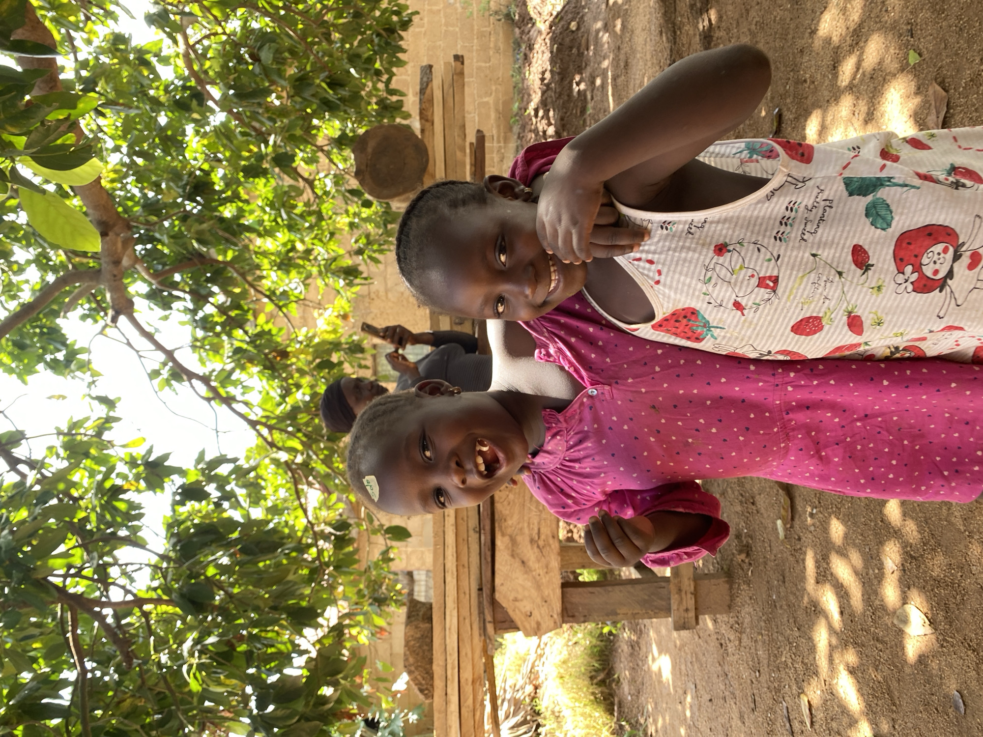 Two smiling young girls are positioned closely together, with the girl in front wearing a pink dress and the girl behind her in a white top decorated with strawberries. They are standing outdoors, with a leafy tree canopy above them and a dirt ground below. In the background, a wooden structure and a person holding a cell phone can be seen. The location is Hoss, Nigeria.