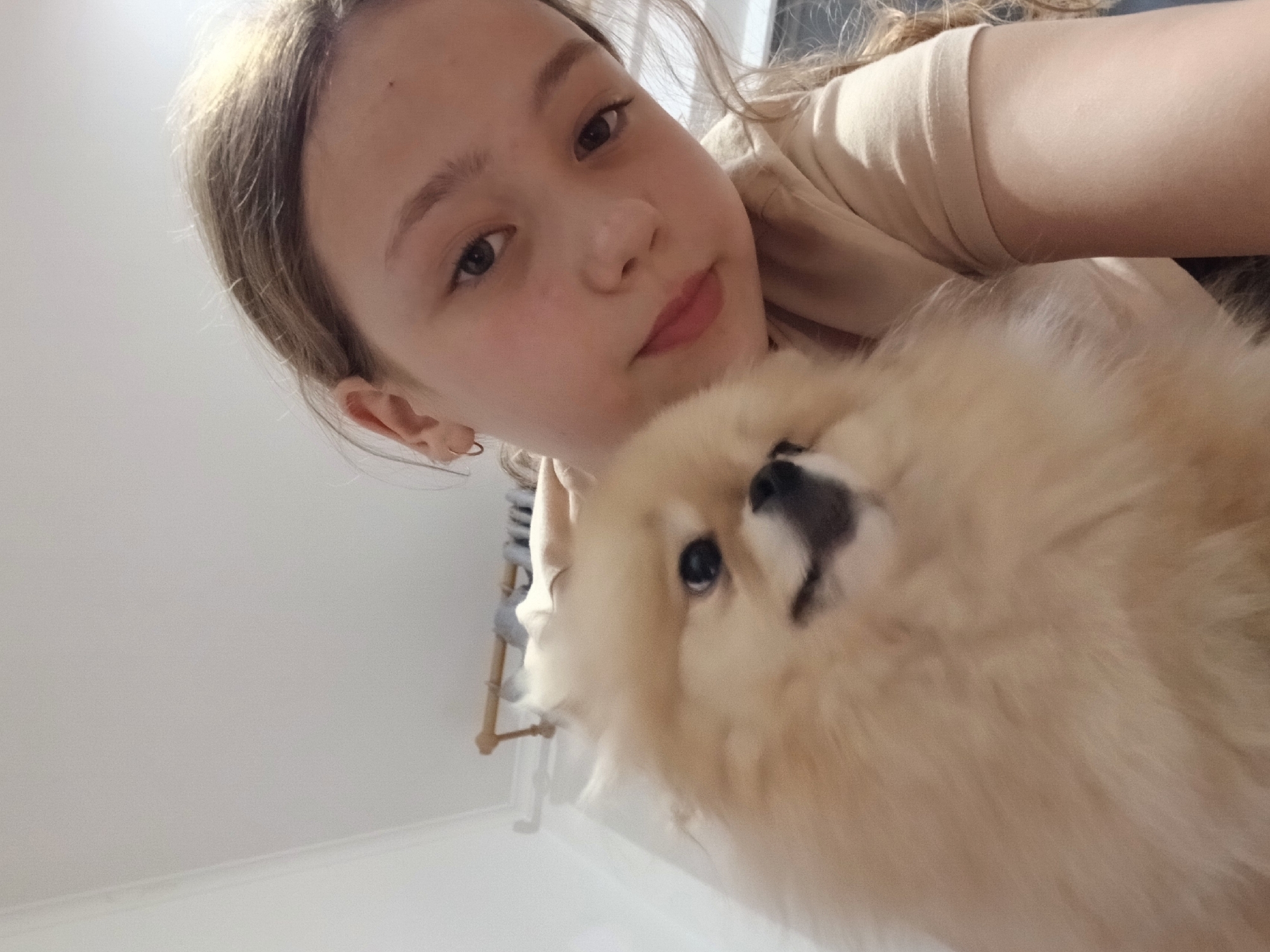 A young girl with light brown hair and fair skin takes a selfie with a small, fluffy, light brown dog. The girl has light eyes, a small nose, and pink lips. She is wearing a beige top. The dog is positioned close to the camera, its black nose and eyes visible. The background appears to be an indoor space with white walls and a clothes rack in the distance. It is impossible to confirm the location as Krasnodar, Russia based on this image alone.