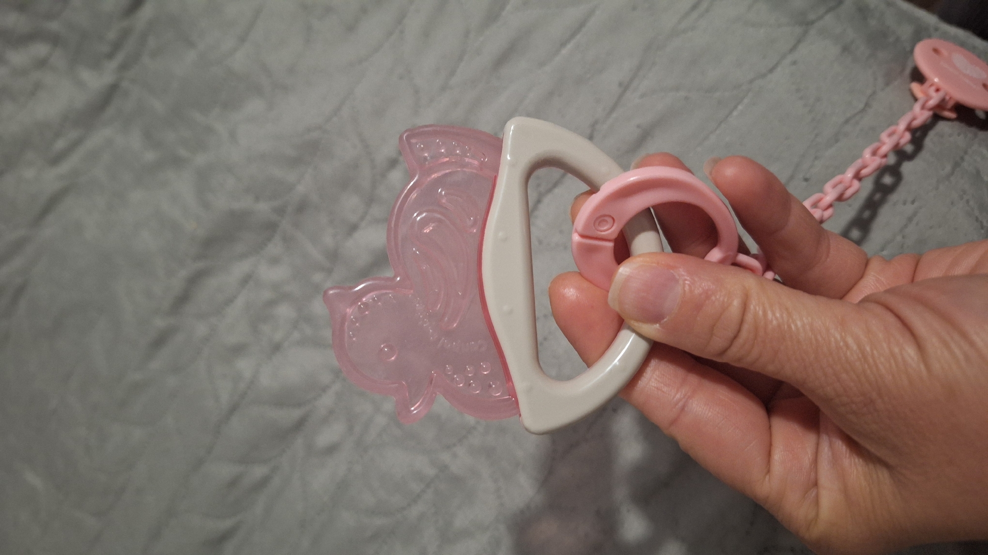 The image captures an adult hand holding a pink, translucent baby teether shaped like a whimsical creature, possibly a unicorn or winged horse with a mermaid-like tail. The teether has a white or light grey handle for gripping. Connected to the hand or the teether assembly is a pink pacifier clip featuring a chain-link design, leading to a circular attachment at its visible end. The background is a soft, grey, textured fabric, likely a blanket or quilt. The lighting indicates an indoor setting, with some reflections on the plastic surfaces. There appears to be some raised, indecipherable text or branding molded into the pink teether. The scene is set in Blagoevgrad, Bulgaria.