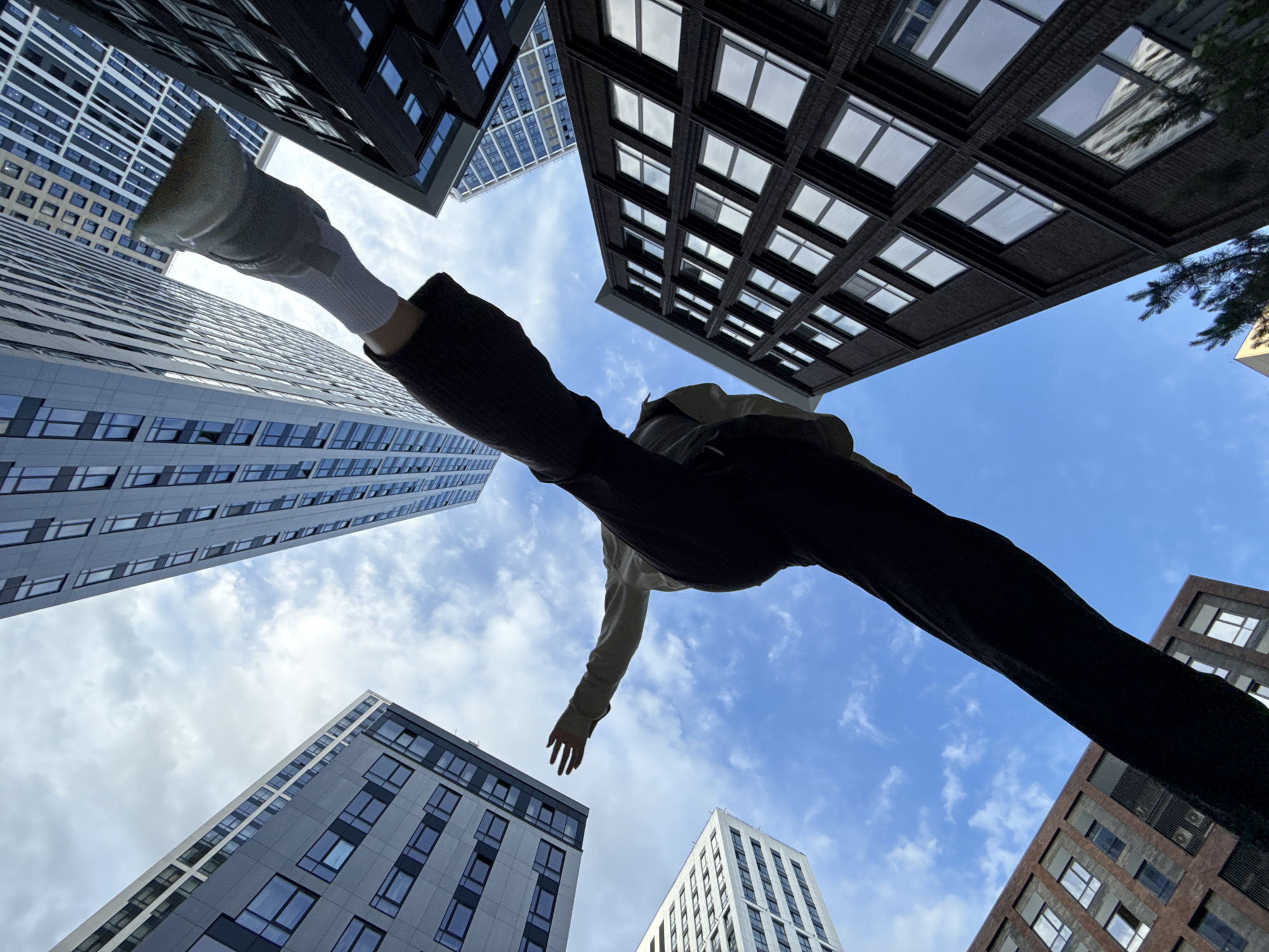 A low-angle shot captures a person's legs and torso extended upwards against a backdrop of modern city architecture and a cloudy sky. The individual is wearing dark pants and a light-colored long-sleeved shirt, with one leg raised high and the other bent. Their arms are outstretched, seemingly balancing themselves. Surrounding the person are tall buildings with geometric window patterns, some dark and brick-faced, others light gray and more contemporary. The perspective emphasizes the height of the structures and the vastness of the sky. The scene appears to be an urban setting, possibly a city square or street, with the architecture suggesting a contemporary design. The time of day is likely daytime, given the presence of natural light and the clear sky with some cloud cover. The overall impression is one of dynamic movement and urban exploration.