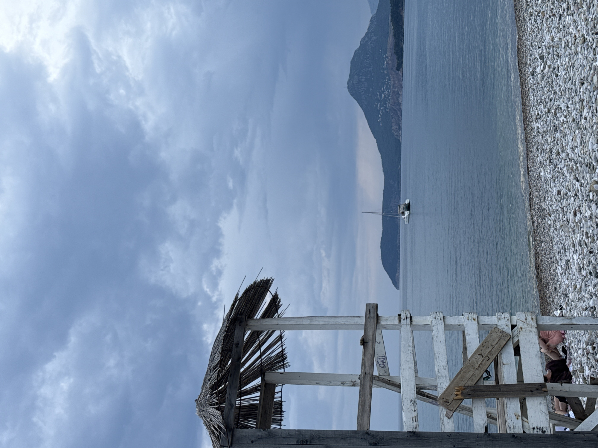 The image depicts a cloudy day at a pebble beach in Bar, Montenegro, with a view of the Adriatic Sea. In the foreground, a weathered wooden lifeguard stand with a thatched roof stands prominently on the left. The stand is partially constructed with white-painted wood and some sections appear damaged or unfinished, with visible nails and rough-hewn planks. Two individuals are seated on the ground behind the stand, partially obscured by its structure, facing towards the sea. One person wears a dark top and the other wears pink shorts. The beach is covered in smooth, grey and white pebbles. The sea is calm and a light blue-grey, with a white catamaran sailing in the middle distance. Across the water, a mountainous landscape with scattered buildings and a prominent peak dominates the horizon. The sky is filled with heavy, grey clouds, suggesting overcast or potentially inclement weather.