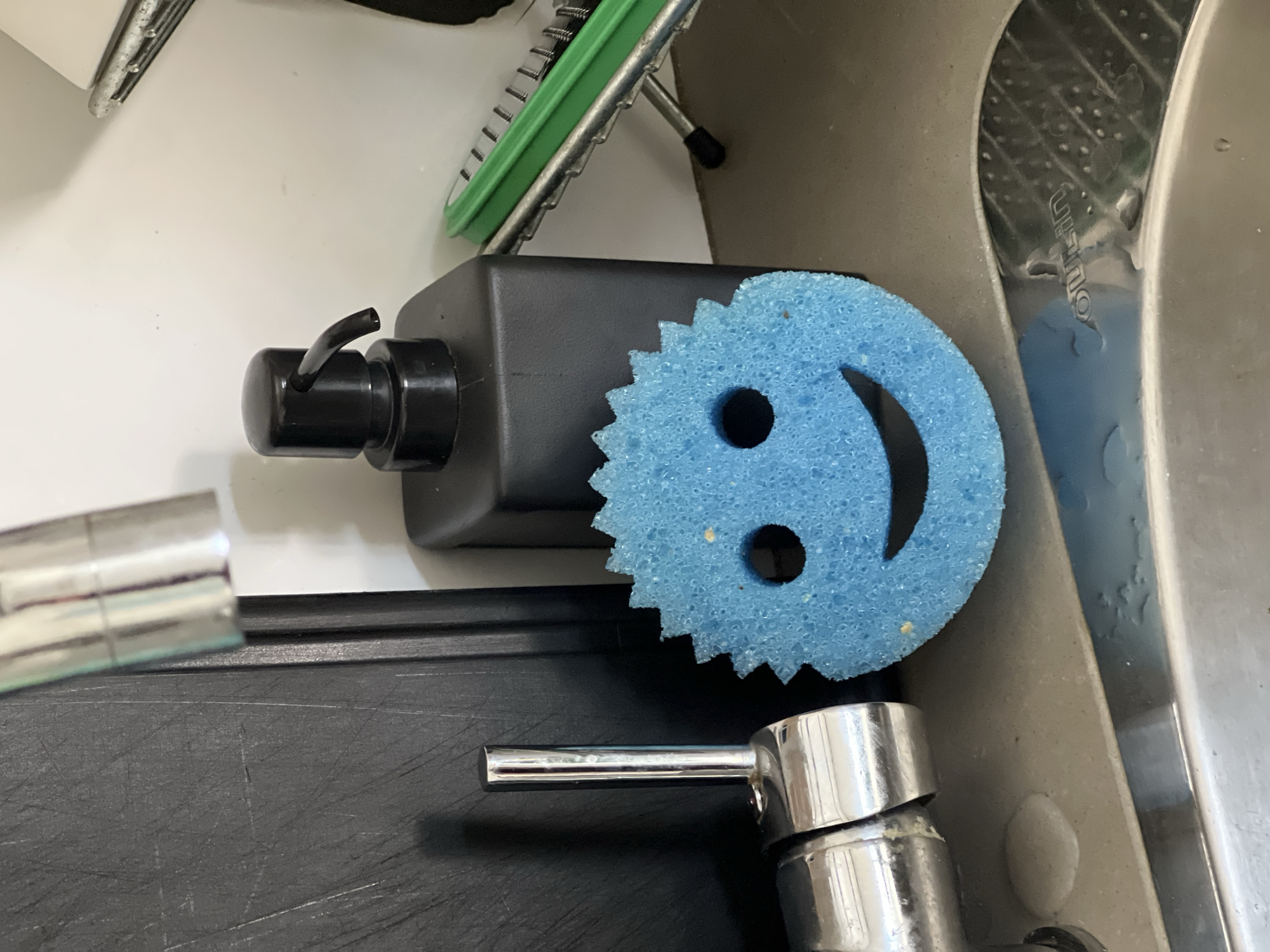 The image presents a close-up, top-down view of a kitchen sink area. Dominating the center is a bright blue, circular scrubbing sponge with a serrated edge, featuring two circular cut-outs for eyes and a curved cut for a mouth, giving it a smiling face appearance. This sponge rests on top of a black, rectangular pump bottle, likely containing dish soap, which is situated on a dark-colored countertop or mat.
To the bottom left, a shiny, chrome-finished faucet or tap extends into the frame. In the upper background, part of a metallic dish drying rack with green plastic accents is visible. The main sink basin, made of a light grey or beige material, occupies the right side of the frame, alongside a section of a stainless steel sink. Water droplets are visible on the sink surfaces and metallic elements, suggesting recent use.
Faint text, appearing to spell "oulilin," is visible on a textured, curved surface within the sink area. The scene is brightly lit, likely by indoor artificial light, and depicts common kitchen items associated with washing dishes. No people or specific activities are shown.