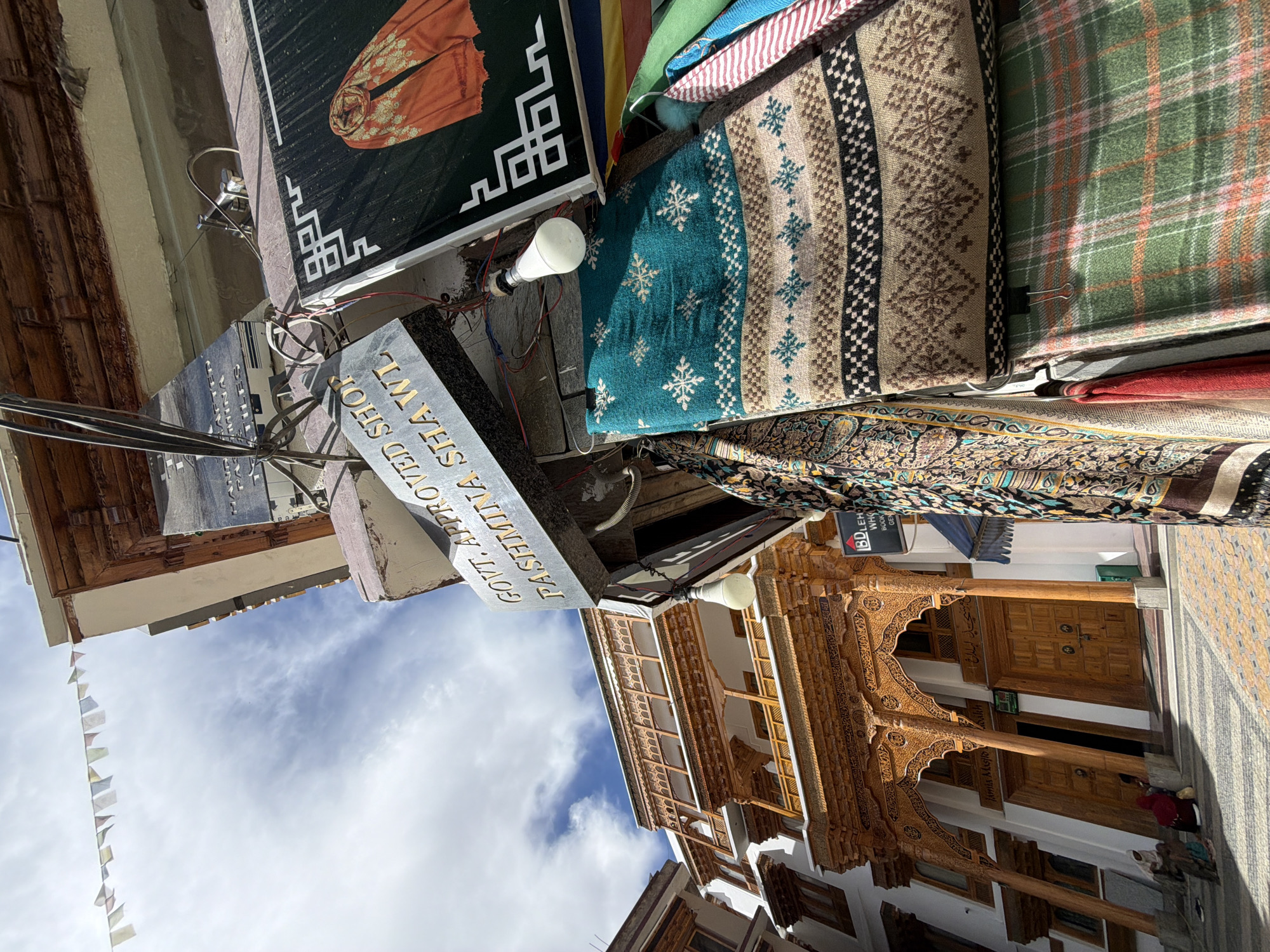 This image captures a street scene in Leh, India, likely near the Jama Masjid Leh. In the foreground, several pashmina shawls are displayed for sale, featuring intricate patterns and vibrant colors, including a teal shawl with snowflake motifs and a green plaid shawl. A sign overhead reads "GOVT. APPROVED SHOP PASHMINA SHAWL." Behind the shawls, the elaborate wooden architecture of a building, possibly the Jama Masjid Leh, is visible, with detailed carvings and balconies. Prayer flags are strung across the sky. A few individuals are seated on the ground in front of the building, appearing to be locals or visitors. The sky is partly cloudy, and the sun is casting shadows. The overall scene depicts a bustling market area with a strong cultural presence.