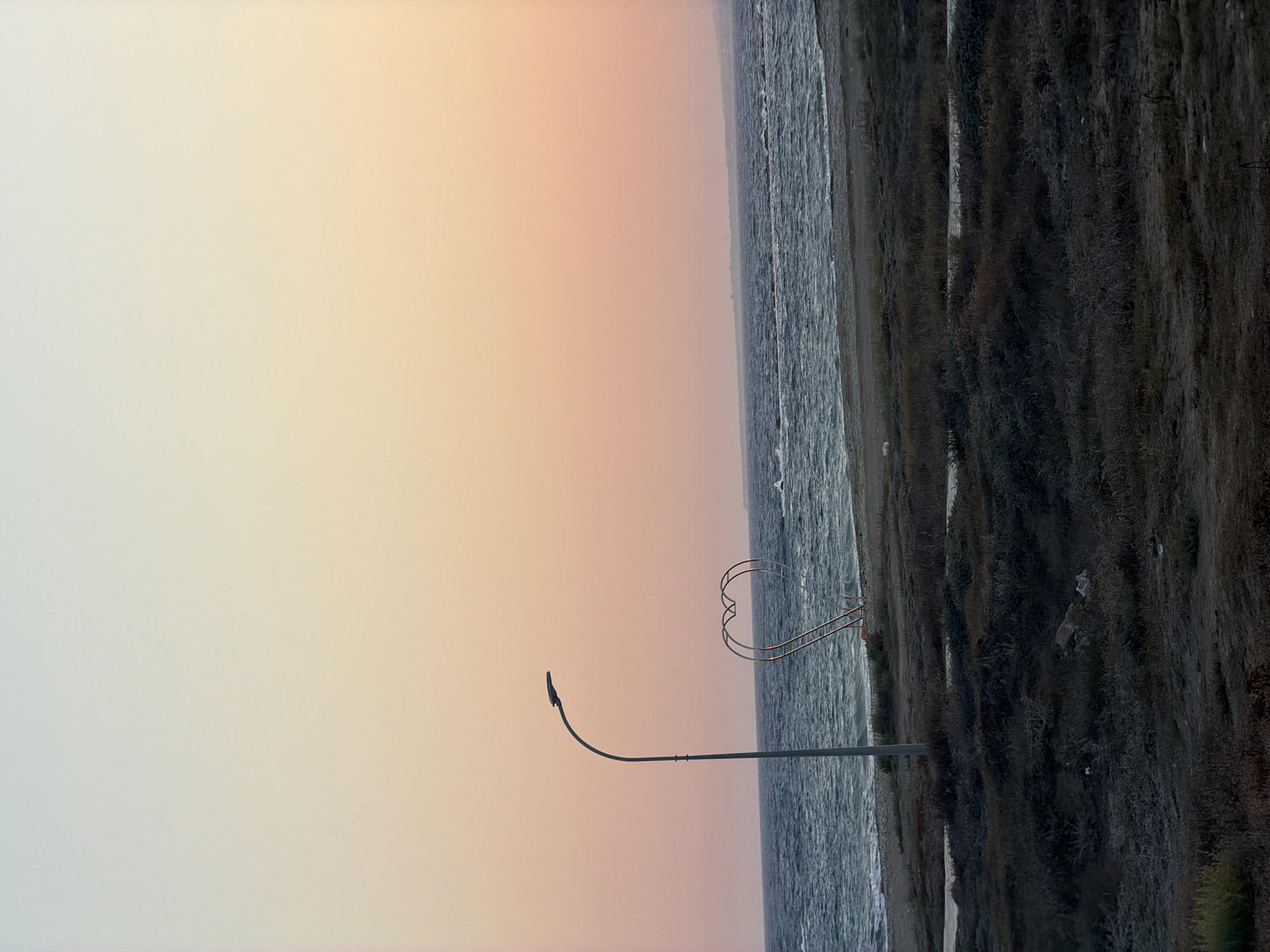 The image depicts a beach scene at Meneou, Cyprus, during what appears to be sunset or sunrise. A tall, grey street lamp stands on the left side of the frame, its curved arm extending towards the right. To the right of the lamp, a large, metallic heart-shaped sculpture is visible on the sandy shore. The sea is in the background, with moderate waves and a hint of whitecaps. The sky exhibits a gradient of colors, transitioning from a pale blue at the top to warm orange and pink hues near the horizon. The foreground consists of dry, sparsely vegetated ground. There are no people clearly visible in the immediate vicinity of the lamp or the heart sculpture, though distant figures or objects might be present on the water or in the far distance.