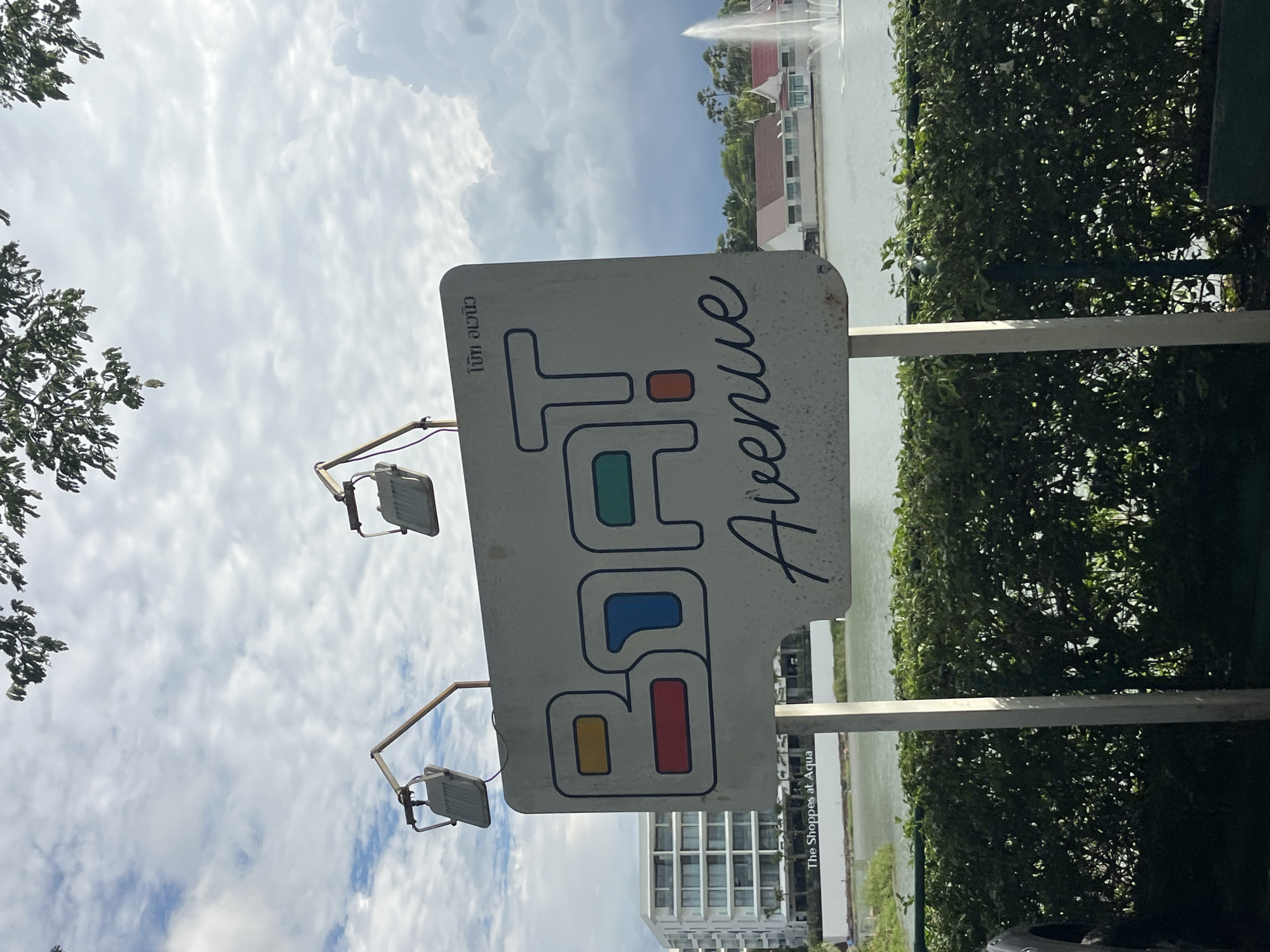 A rectangular sign with rounded corners, mounted on two metal posts, stands in front of a body of water. The sign has a white background and features the text "BOAT Avenue" in large, stylized letters. The letters "B", "O", "A", and "T" are outlined in blue and filled with different colored rectangles: yellow, red, blue, and green respectively. The word "Avenue" is written in a cursive, dark blue font below the block letters. In the upper right corner of the sign, there is Thai text. Two floodlights are attached to metal arms extending from the top of the signposts, angled downwards.
In the background, to the left, is a modern, multi-story building with balconies. Below the building, the text "The Shoppes at Aqua" is visible on a white wall. Across the water, there are several buildings with red roofs and white walls. A fountain with a tall plume of water is visible in the far right background. The sky is cloudy with patches of blue. Lush green bushes line the foreground, partially obscuring the base of the signposts. The overall scene appears to be an outdoor commercial or residential area, likely a waterfront development. The sign is a prominent feature, identifying the "BOAT Avenue" location within the city of Choeng Thale, Thailand.