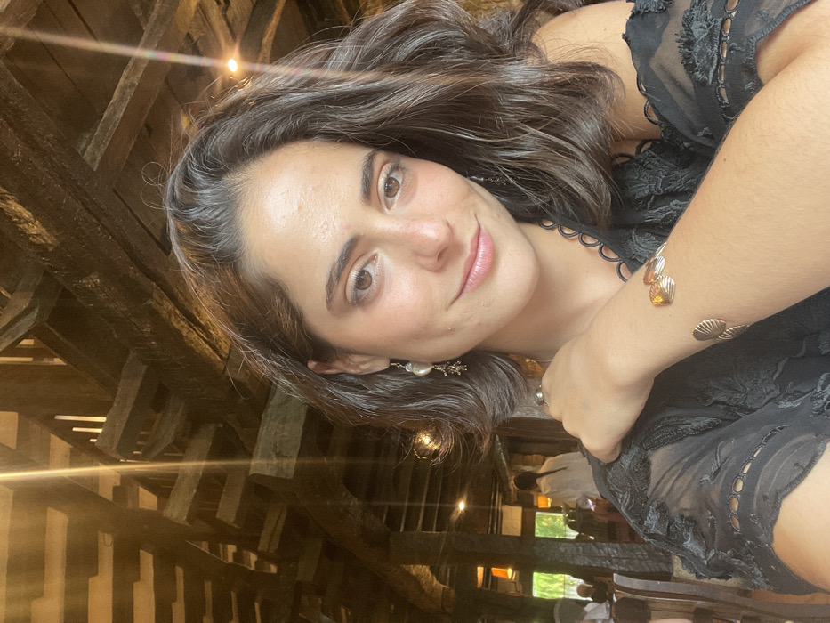 An adult female is depicted in a portrait-style photograph taken inside Restaurante Zelaieta in Amorebieta-Etxano, Spain. The location is identifiable by its distinctive interior architecture, featuring a dense network of heavy, dark wooden beams forming the ceiling and structural elements of the dining area. The female subject, positioned in the foreground, has medium-brown skin, dark brown, wavy hair, and brown eyes, and is looking directly at the camera with a subtle smile. She is wearing a dark, possibly black, semi-sheer garment with patterned sleeves. Accessories include drop earrings with a pearl-like component on her left ear, a ring on her right ring finger, and a gold-toned cuff bracelet adorned with multiple shell-shaped motifs on her right forearm. Her right hand is visible, resting near her left shoulder. In the blurred background, other patrons are seated at tables in the dimly lit environment, indicating a public dining setting.
