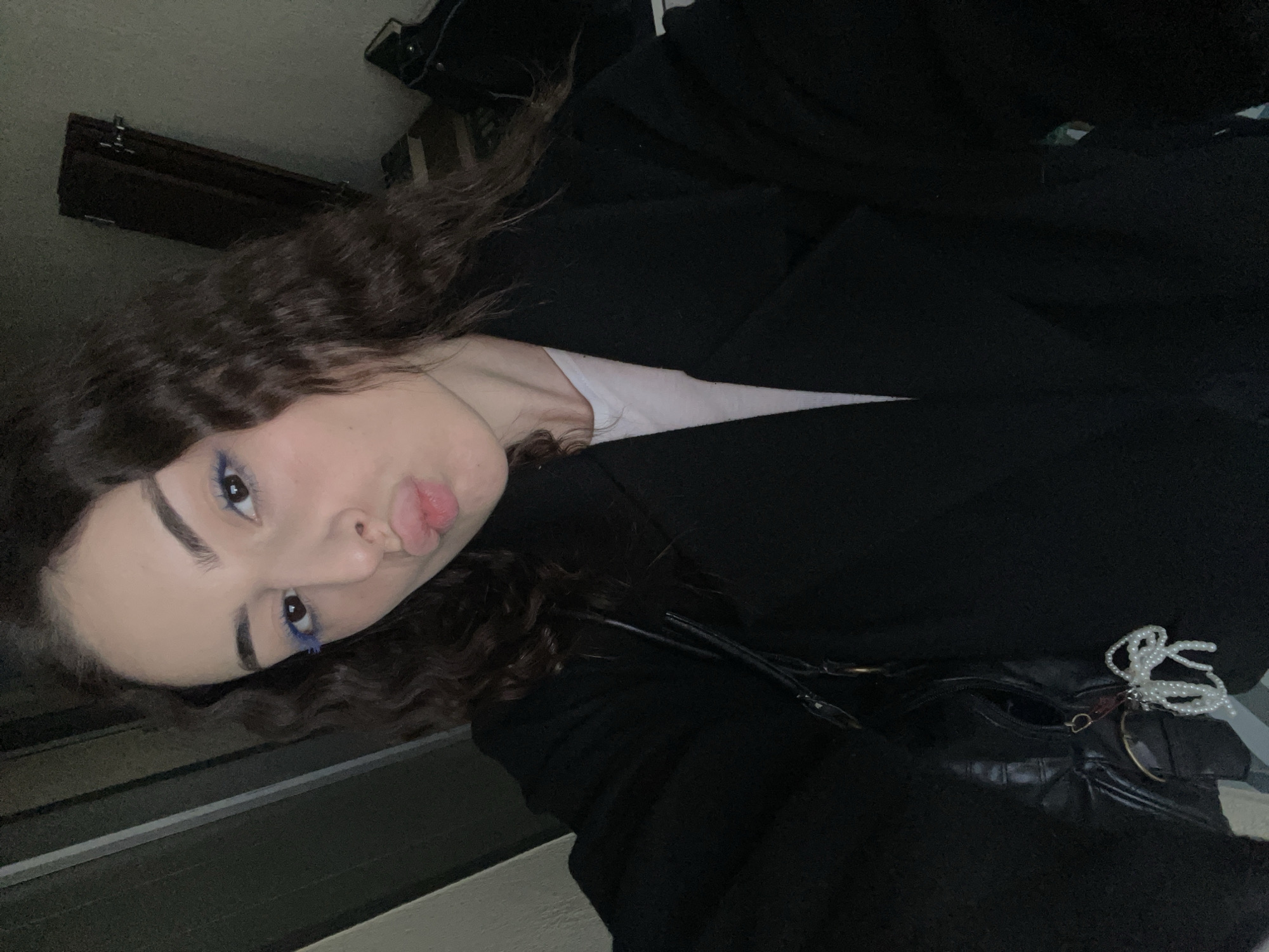 A young woman with curly hair and blue eyeshadow is lying down, puckering her lips towards the camera. She is wearing a white shirt and a black blazer. A black purse is visible. The location cannot be determined as Moscow, Russia.