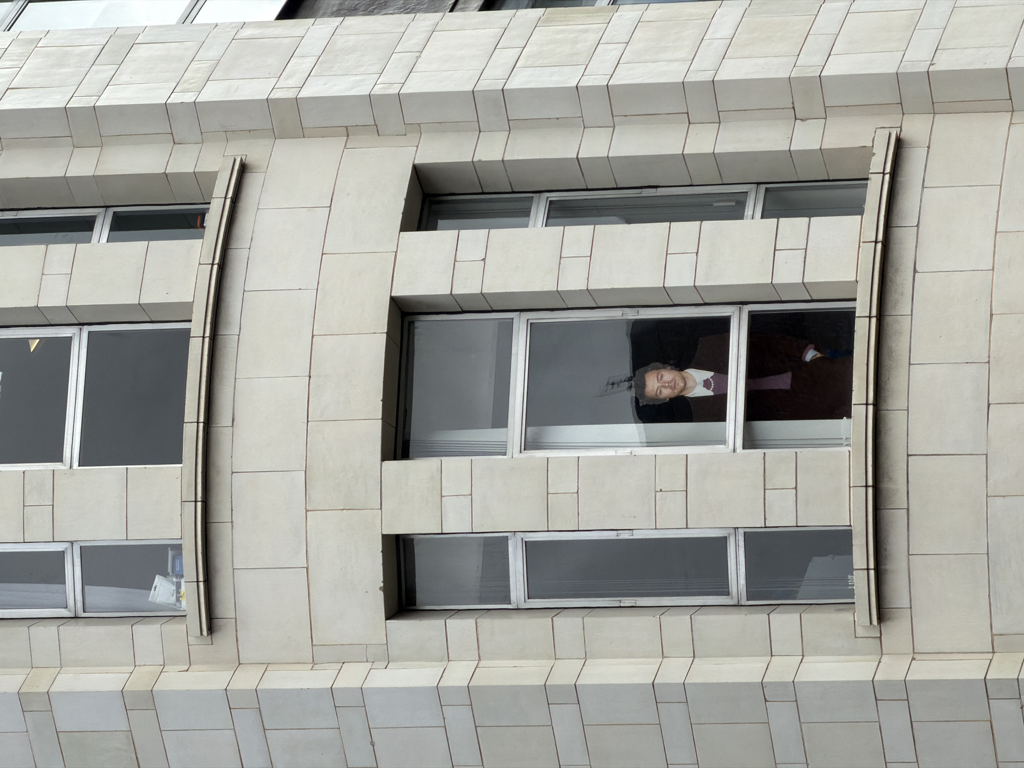 A medium, straight-on shot shows part of a light gray building exterior with rectangular stones, some of which are layered in a staggered fashion. On the side are long, vertically grooved bars. The building features windows with grayed-out panes. One of the windows has a person's reflection inside. The person is looking at the viewer and appears to be a man in a brown or maroon jacket.