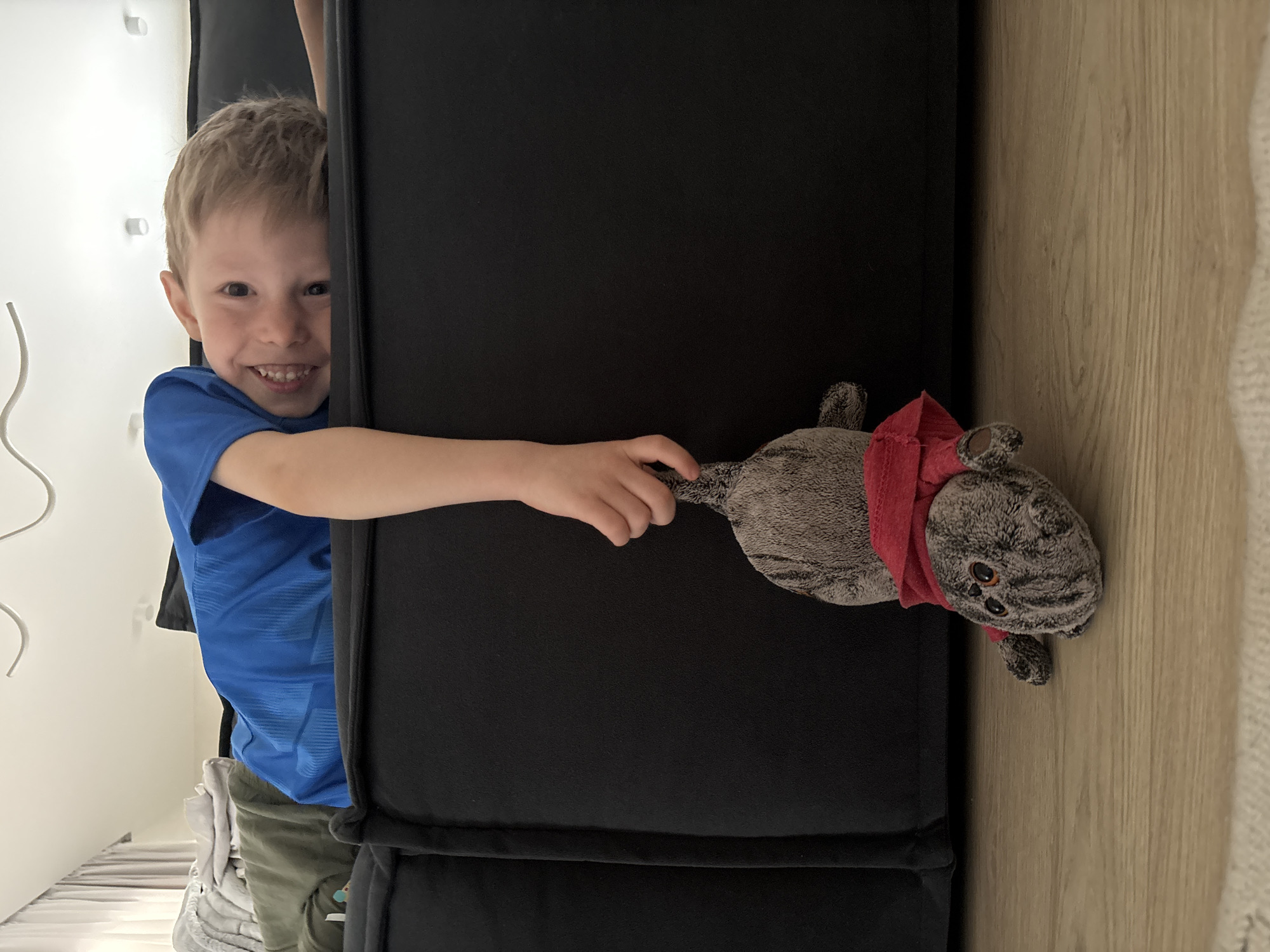 The media file displays a playful indoor scene, rotated 90 degrees clockwise from a typical upright orientation, located in Chaikovsky, Russia. A young boy, with light hair and a bright, toothy smile, is peeking out from behind a large, dark-colored, vertical panel, possibly part of a cabinet or furniture. He is wearing a blue t-shirt and olive-green shorts, with white sneakers visible near the bottom left.
The boy's right arm extends horizontally across the dark panel, holding a grey, striped plush toy by its tail. The toy, which resembles a cat and wears a red scarf, dangles to the right. On the far left of the image, behind the boy's head, is a light-colored wall with faint vertical elements and a white wavy cord. To the far right, beyond the dark panel and the toy, is a light-toned wooden wall with a distinct grain. The scene is well-lit, suggesting daytime. The boy's clear expression of happiness and the playful action with the toy are prominent. No text is visible in the image.