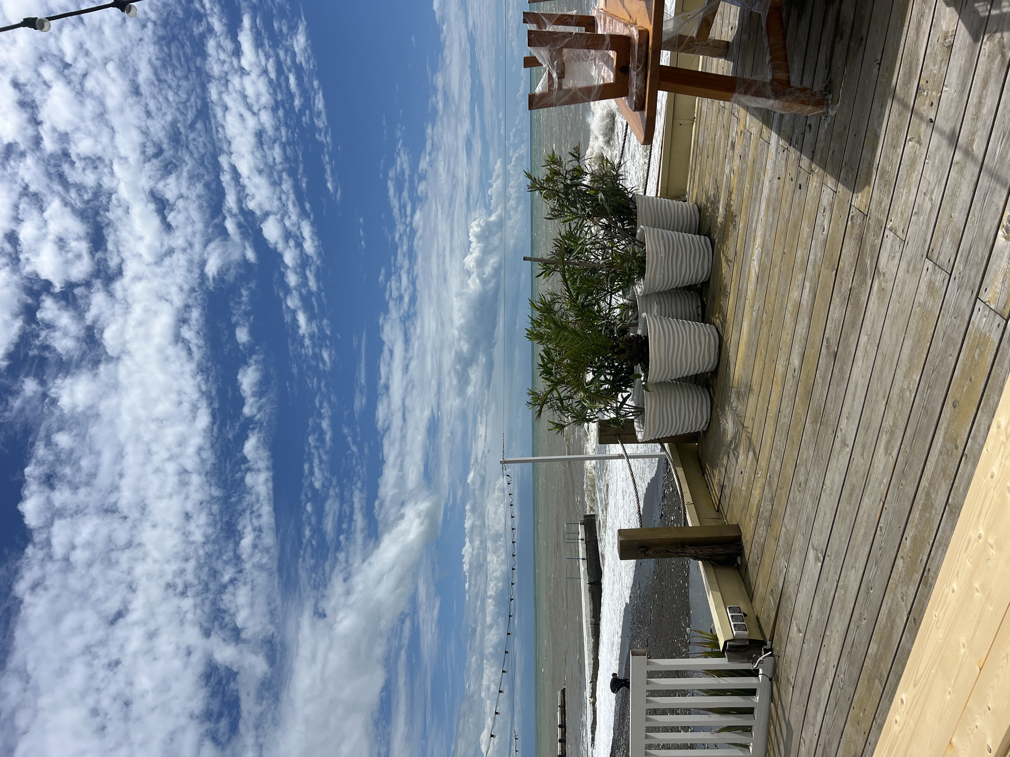 A partly cloudy sky is visible above the Black Sea in the city of Sochi, Russia. The water is a dull green-gray color, while the sky is bright blue. There are a few white clouds in the distance. A wooden deck with a white railing is visible in the foreground. There are three white, ridged planters on the deck, each containing green plants. A wooden table and chairs are also on the deck. The chairs are partially covered in clear plastic. A string of lights is strung along the edge of the deck.