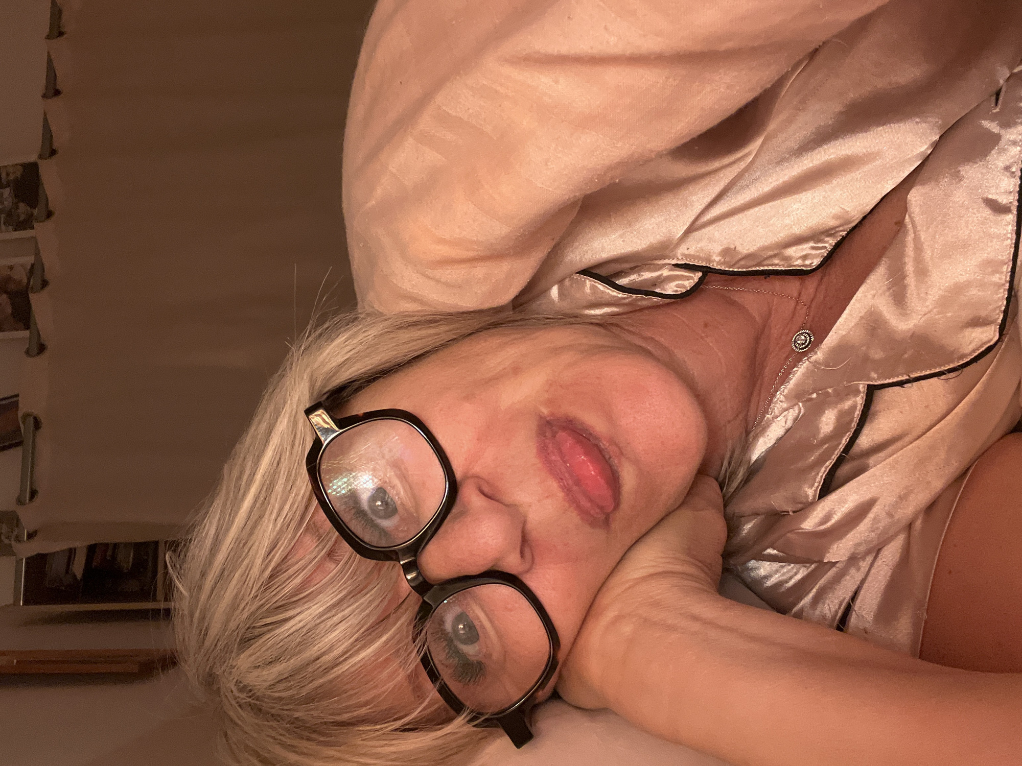A female individual is depicted in a close-up, horizontal orientation, appearing to be lying down in an indoor setting in Chambœuf, France. She is fair-skinned, with short blonde or grey hair and bangs. The individual wears dark-framed eyeglasses and a light-colored, possibly champagne or rose gold, satin-like top with black piping, resembling pajamas. A silver-toned chain necklace with a small, circular, possibly diamond-set pendant is visible around her neck. Her left hand supports her head, which rests on a light-colored surface, likely a pillow. She is engaged in an expressive action, with her eyes wide open, pupils dilated, and her tongue extended out from her mouth. A peach or pink-colored blanket or duvet is pulled up around her right shoulder. The background is dimly lit and out of focus, revealing a curtain rod with several rings and indistinct framed objects (likely pictures) mounted on a wall above it. The overall lighting is warm and artificial.