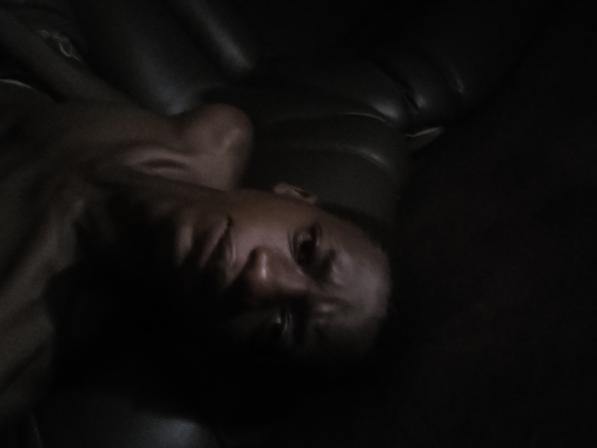 The image depicts a dimly lit interior. A person with dark skin lies on what appears to be a dark leather surface, possibly a couch or bed. The individual's face is visible, with the features partially obscured by shadow. The exact location in the city of Unknown, Unknown cannot be determined from the image.