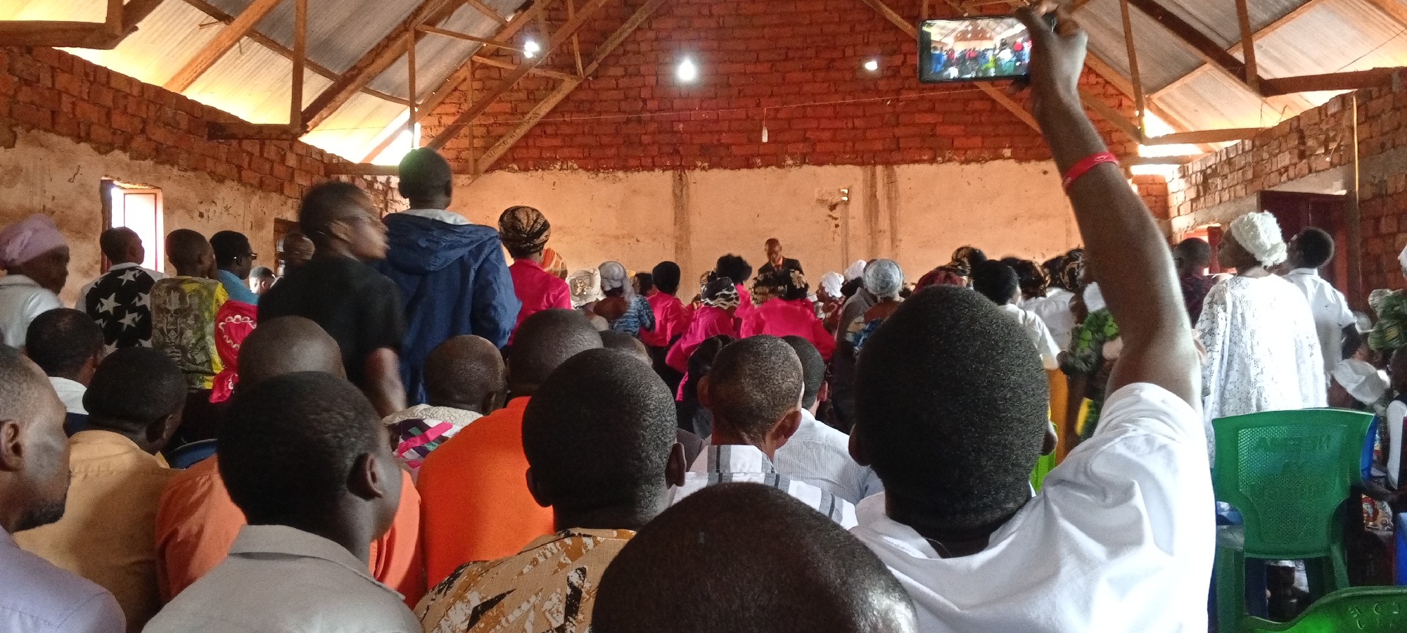 Captured in Ngalula, Democratic Republic of the Congo, the scene shows an assembly of people indoors, likely at a gathering or meeting. The subjects are predominantly Black individuals. The room has a brick interior with a roof structure visible. One person is holding up a mobile phone, possibly recording the event or streaming it live. The room is filled with individuals seated or standing, with their backs to the camera. People are wearing various types of clothing, including pink shirts, white dresses, and colorful patterned fabrics. There is a chair in the foreground on the right. The overall environment suggests a community event taking place in a simple, brick-structured building.