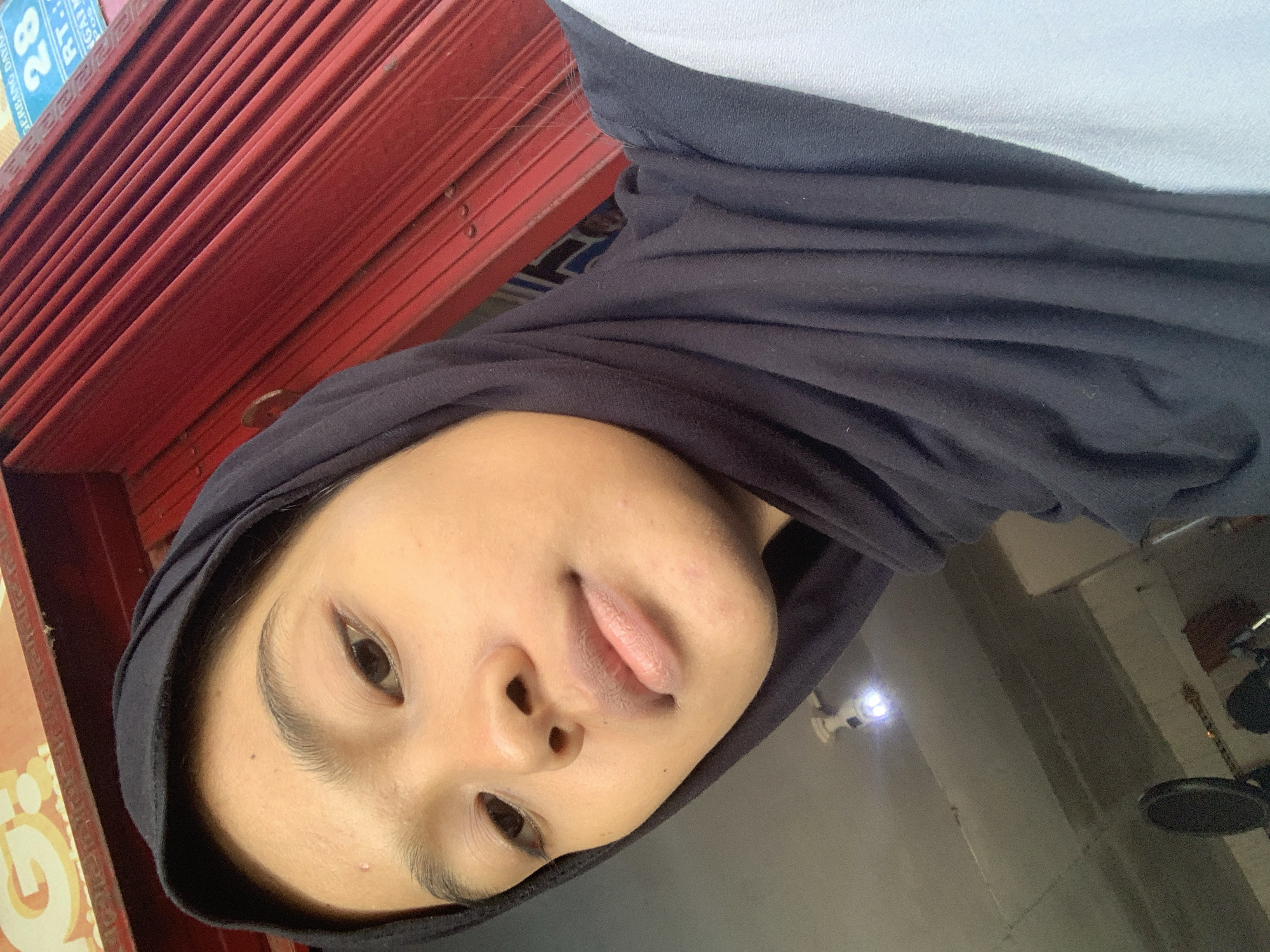 In Sungai Meriam, Indonesia, a young woman wearing a black hijab and a light blue garment is captured in a close-up, front-facing selfie. Her skin is lightly tanned, and she has distinct eyebrows and eyes. The background features a red corrugated metal shutter with some signs visible, and a ceiling with a light fixture and what appears to be soundproofing material or acoustic panels. The overall lighting suggests an indoor environment.
