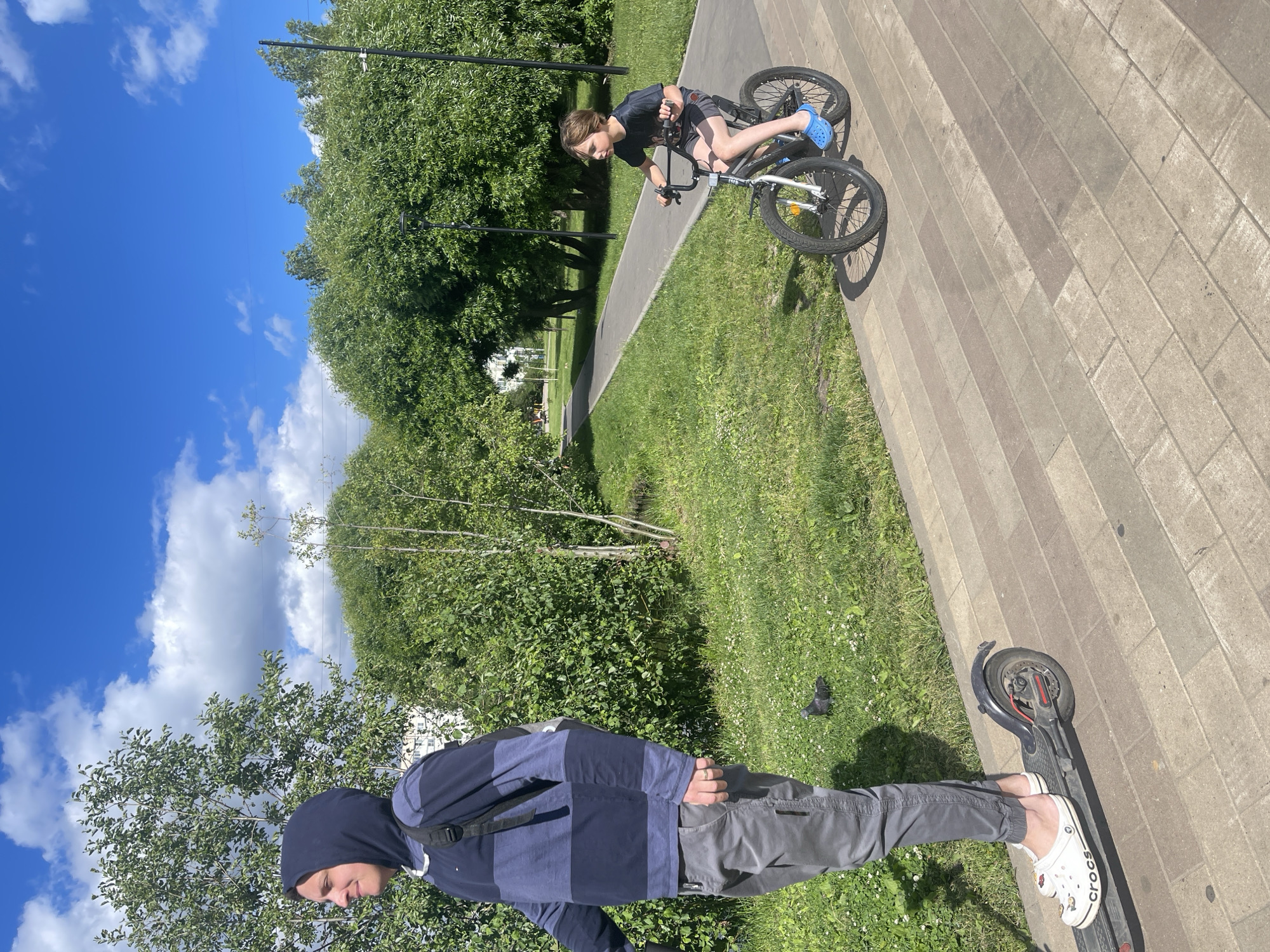 On a bright, sunny day, two young males are engaged in outdoor activities in a park or recreational area. On the left, a person wearing a dark blue and navy striped hooded sweatshirt and grey pants stands on a black electric scooter, facing left. They are wearing white Crocs sandals. To the right, a boy wearing a dark t-shirt and grey shorts rides a bicycle. He is wearing blue Crocs sandals. The path they are on appears to be paved with reddish-brown and grey rectangular bricks, bordered by a grassy area with scattered white flowers. Lush green trees and bushes form the background, with a clear blue sky with scattered white clouds above. The setting is consistent with a city park, possibly in Moscow, Russia, given the presence of manicured landscaping and paved pathways.