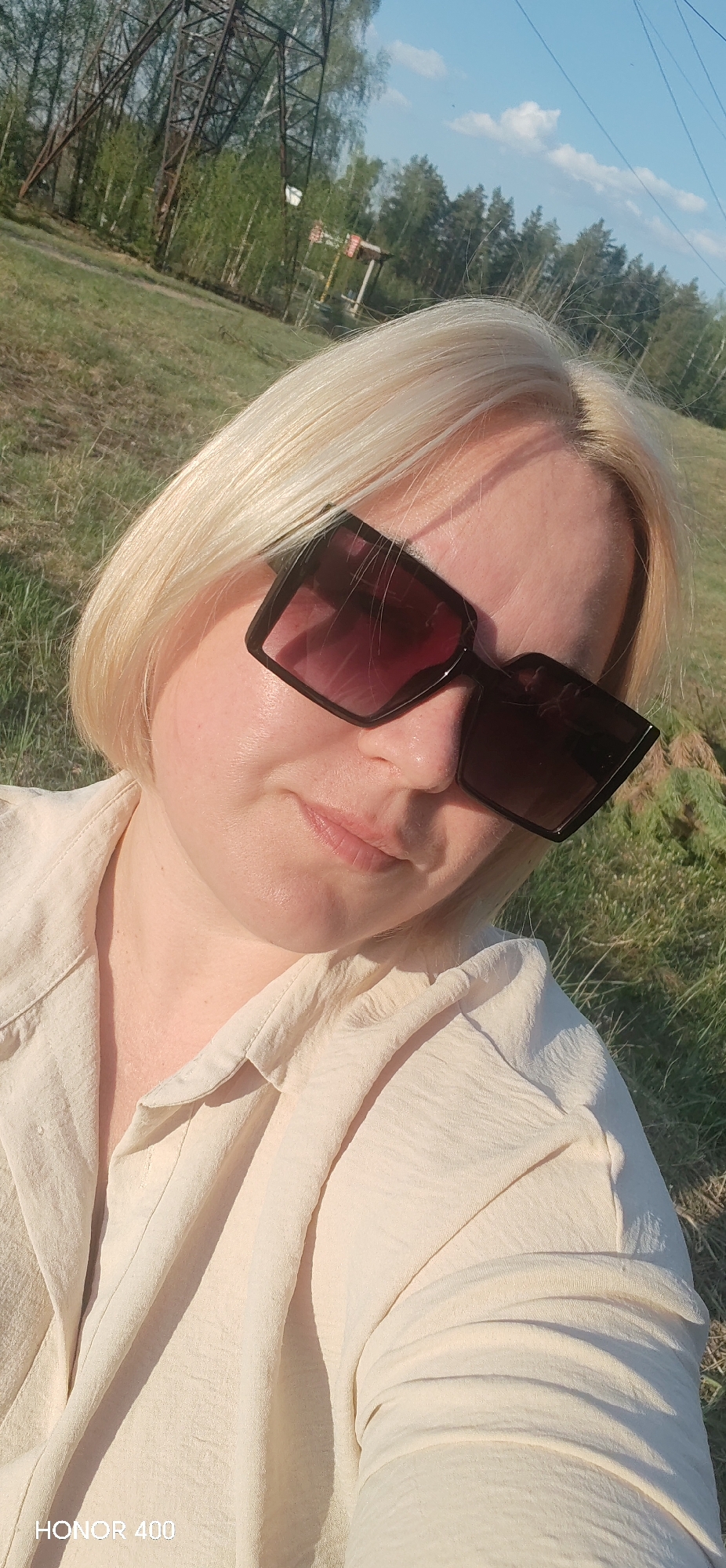 A woman with fair skin and blonde hair is wearing dark sunglasses and a light-colored collared shirt. She is outdoors in a grassy area near a forest. A transmission tower is visible in the background. The image was taken by an Honor 400, likely in Noviy, Russia, based on geographical clues in the image's context.