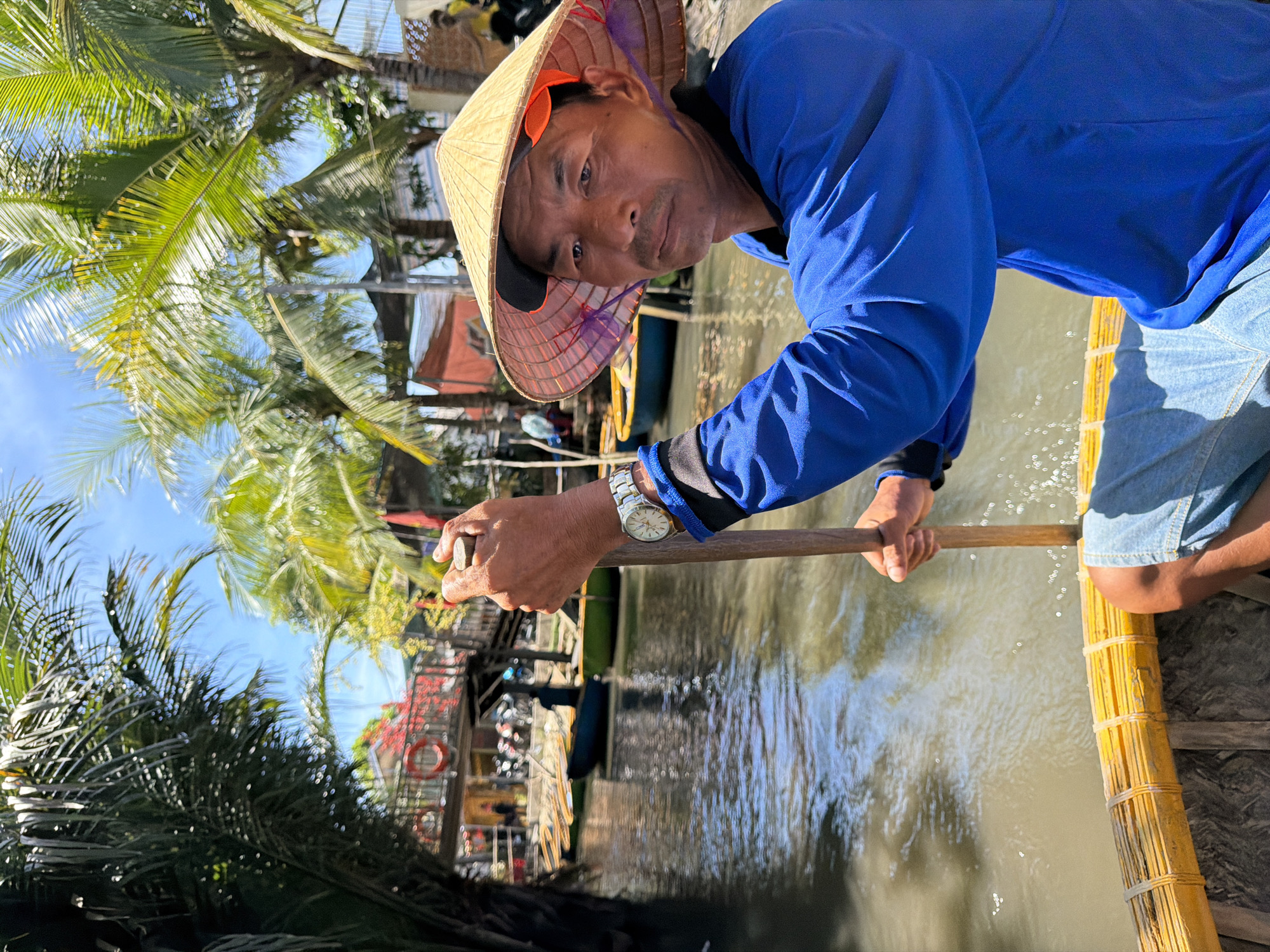 The image depicts a man wearing a conical hat and a blue shirt, holding a wooden oar while sitting in a boat on a canal in Hoi An, Vietnam. He is looking towards the camera with a serious expression. The boat appears to be a traditional basket boat, made of woven bamboo. The canal is lined with palm trees and buildings with red roofs, suggesting a tropical environment. In the background, other boats are visible, and people are walking along the canal bank. The water is murky and reflects the sunlight, creating a shimmering effect. The overall scene captures a moment of daily life in a Vietnamese riverside community.