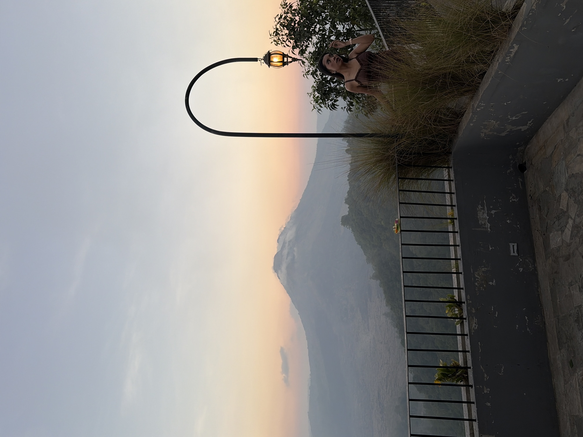 A woman stands on a balcony overlooking a mountain landscape at sunset in Kintamani, Indonesia. The sky is a gradient of pale blue to soft orange, with a prominent volcano shrouded in mist in the distance. A black metal railing runs along the edge of the balcony, with tall, dried grass growing in planters in front of it. A black lamp post with an illuminated orange lantern arches over the scene. The woman, wearing a brown slip dress, is leaning against the railing and looking towards the mountains. The balcony floor is made of stone tiles.