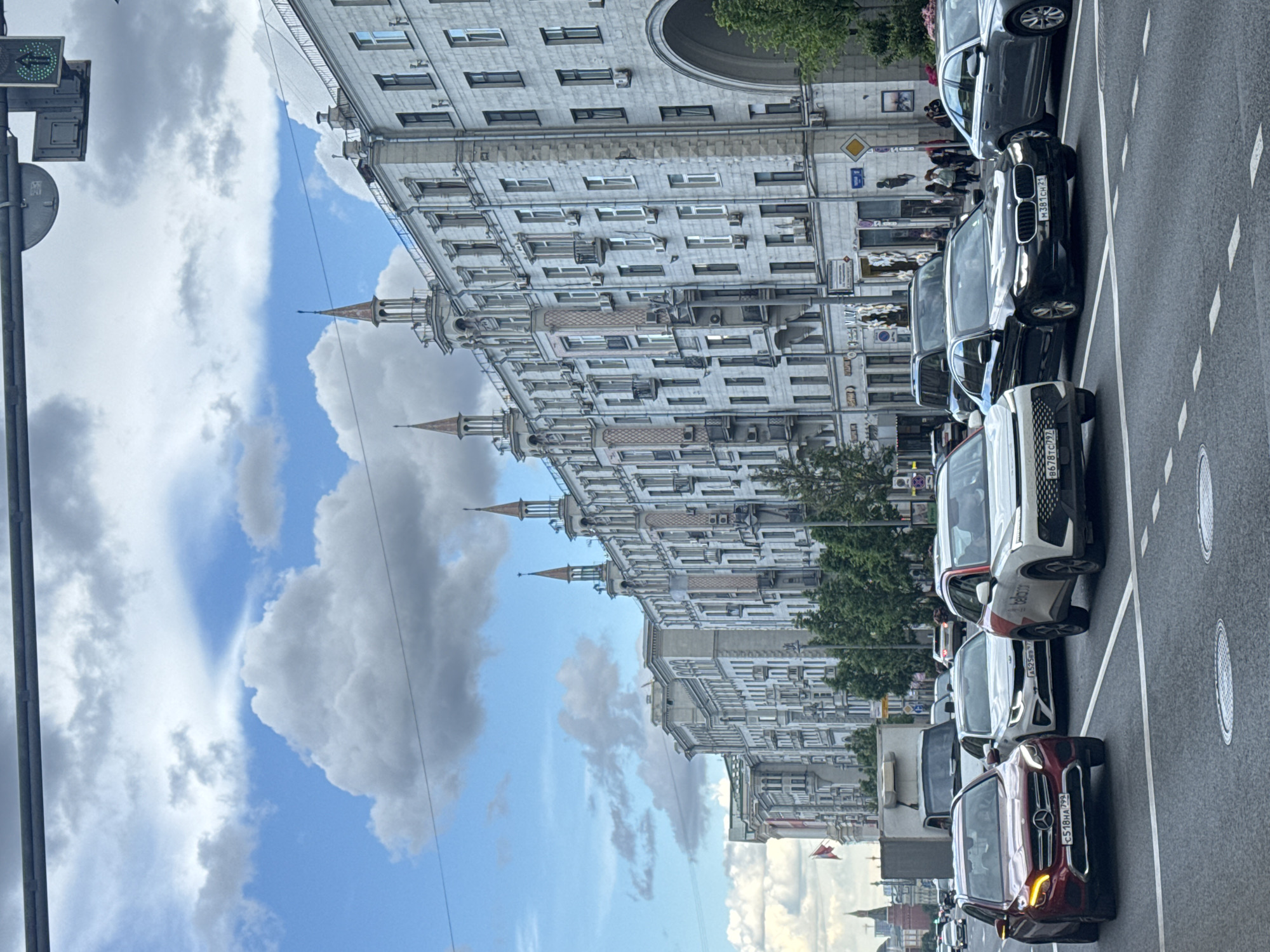 The image depicts a street scene in Moscow, Russia. A row of parked cars lines the right side of the street, with buildings visible on the left side of the street. The buildings are ornate, with multiple stories and architectural details, including towers and arched windows. The sky above features a mix of blue and cloudy sky. There are people walking in the background and traffic lights in the left side.