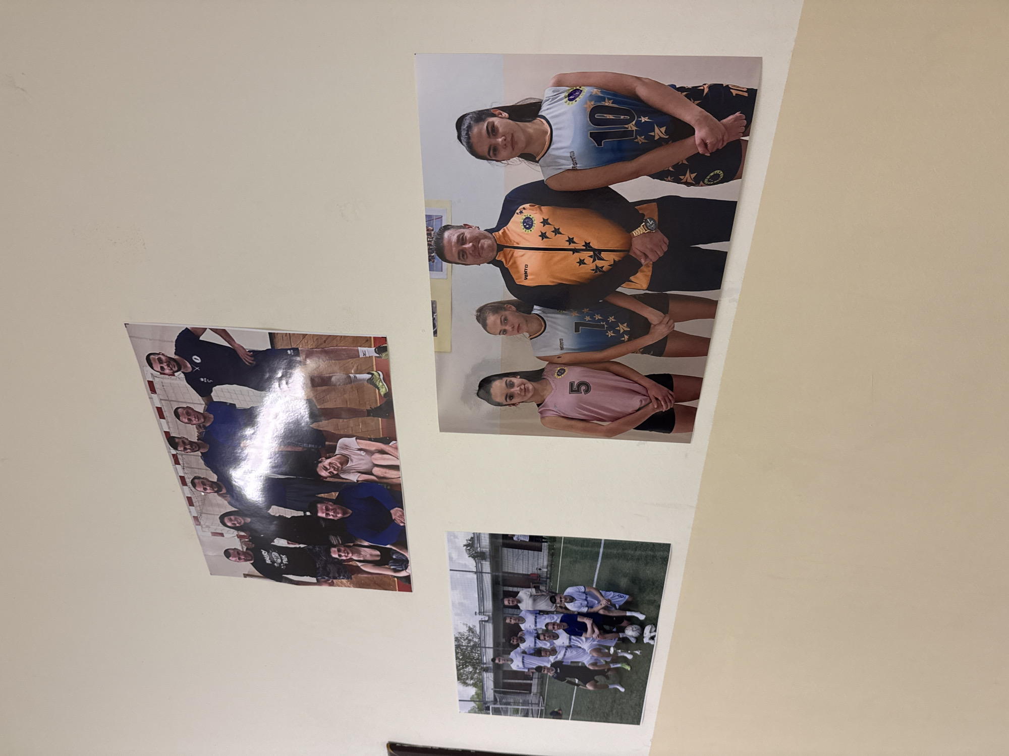 The image displays a section of an interior wall, likely within a sports facility in Sofia, Bulgaria, on which three large photographs are mounted.
The top photograph is a landscape-oriented group portrait of nine adults, composed of six males and three females, posed in an indoor setting featuring a red and white striped goal net in the background. The individuals are dressed in casual athletic wear, including t-shirts and shorts, with one male wearing a shirt displaying "ANGRY BIRDS." All subjects are smiling.
The bottom-left photograph is a landscape-oriented group portrait of ten young adult males on an outdoor artificial turf football pitch. They are uniformly dressed in football kits, with most wearing white jerseys, and some in black or blue. A silver trophy cup and a white soccer ball are positioned on the ground in front of five kneeling individuals. A chain-link fence and structures are visible in the background.
The bottom-right photograph is a portrait-oriented group image of four individuals: one adult male and three young women. The male, positioned centrally, wears an orange and navy blue athletic jacket adorned with star patterns and the "VENTO" brand logo, and a gold-toned wristwatch. To his left and right stand three young women, each wearing a sleeveless athletic top with star designs and numerical identifiers (5, 7, and 10, from left to right) and dark shorts. All four subjects stand with their hands clasped in front of them.
The wall has a light off-white tone in the upper section and a lighter beige/cream tone in the lower section, indicating a subtle color transition.