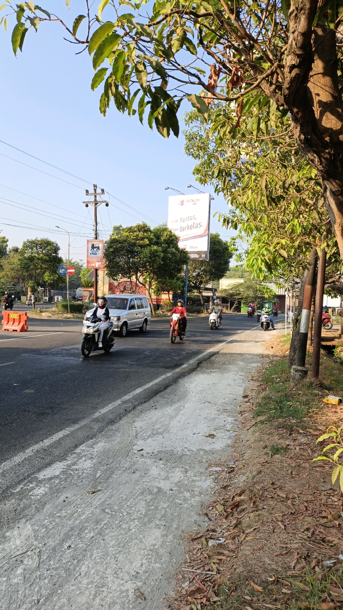 This is a medium-shot, eye-level, vertical, outdoor photograph taken during the daytime in Sumbring, Indonesia. The scene depicts a road with several vehicles and motorcyclists. The road surface is asphalt with a concrete shoulder. On the left side of the road, there are orange traffic barriers and a street light. In the background, there are trees, utility poles, and a billboard.
Specifically, there is a silver van driving on the road. Two motorcyclists are on the road in the foreground. One motorcyclist is wearing a white outfit and riding a white and black motorcycle. The other motorcyclist is wearing a red shirt and riding a red motorcycle. There are several other motorcyclists further down the road. The sky is clear and blue. The foliage of trees is visible in the foreground and background, with some branches extending into the upper part of the frame. The lighting suggests it is either early morning or late afternoon, with shadows cast by the trees.