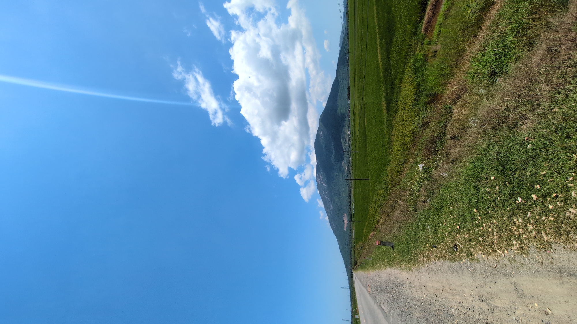 The image depicts a rural landscape in Ninh Hòa, Vietnam, under a clear blue sky with scattered white cumulus clouds. A distinct diagonal lens flare is visible in the upper right quadrant of the sky. In the foreground, a two-lane paved road with a dirt shoulder extends into the distance on the left. White roadside marker posts with orange tops are spaced along the right edge of the road's shoulder. To the right of the road and its shoulder, extensive green agricultural fields, consistent with rice paddies, stretch towards a distant, large mountain range. The mountains appear densely vegetated with dark green foliage, exhibiting lighter patches near the summit. Multiple utility poles with power lines are visible traversing the fields and along the distant left side of the road. No individuals, animals, or vehicles are present in the scene. Minor debris is scattered along the grassy verge adjacent to the road.