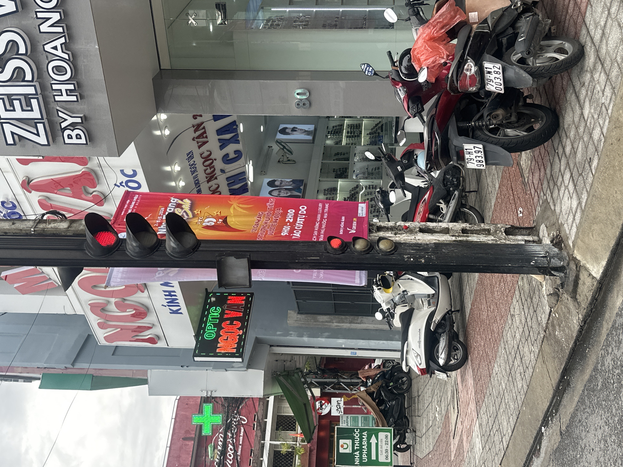 The scene depicts a street in Nha Trang Ward, Vietnam, with various commercial establishments. A traffic light pole is visible, displaying red lights. Adjacent to it are signs for "ZEISS BY HOANG," "KÍNH NGỌC VÂN," and "OPTIC NGỌC VÂN." A red cross sign indicates a pharmacy. Parked motorcycles are present on the sidewalk, with license plates visible on some. Additional signs advertise promotions for "NGỌC VÂN" and "NHÀ THUỐC UPHARMA." Building number 80 is visible.