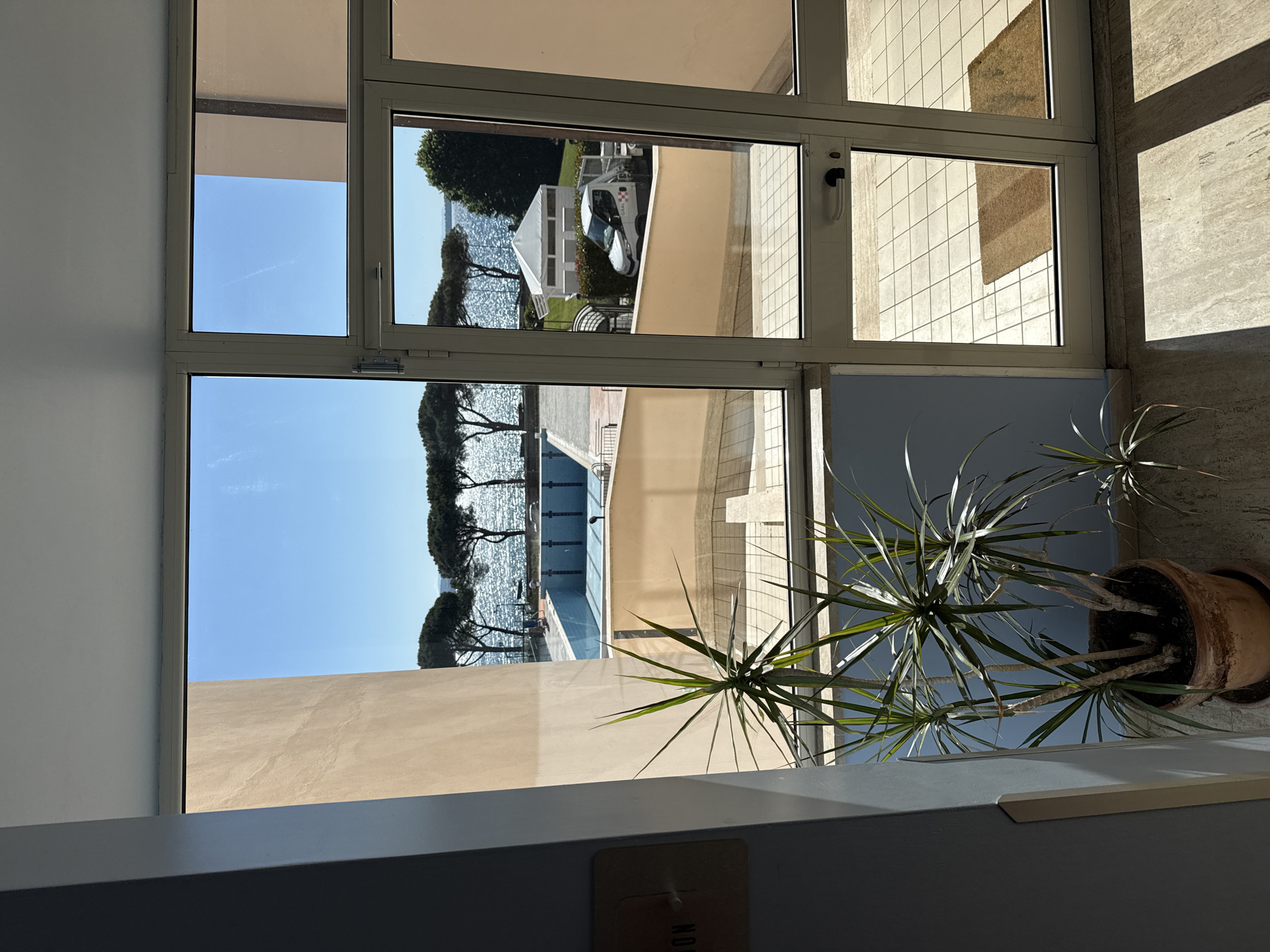 A view from inside a building looks out through a large window at a lake and surrounding property. The scene is dominated by the blue sky and lake water. Trees are visible along the water's edge. Adjacent to the lake is a swimming pool and building. The window frame is visible. A potted plant sits indoors near the window. The location is Bracciano, Italy.
