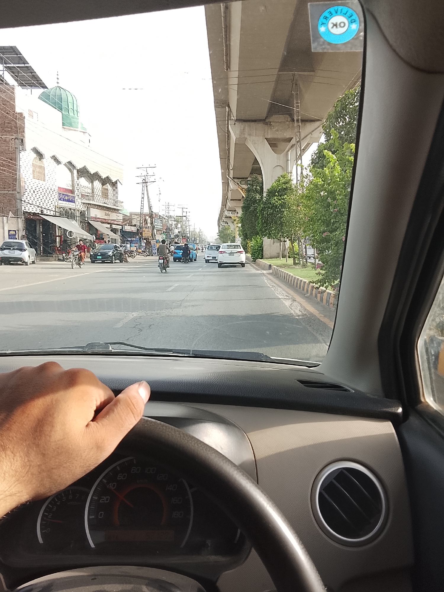 The view from inside a car moving along a road in Gulgasht Colony, Pakistan. The driver's hand rests on the steering wheel, with the speedometer visible. The car is traveling on a road with multiple lanes in each direction. Other cars and motorcycles share the road, moving in both directions. Buildings line the left side of the street, including one with a green dome, likely a mosque. Trees line the right side of the street. A flyover structure is visible above the road. A round sticker is visible on the windshield. The lighting suggests it is daytime.
