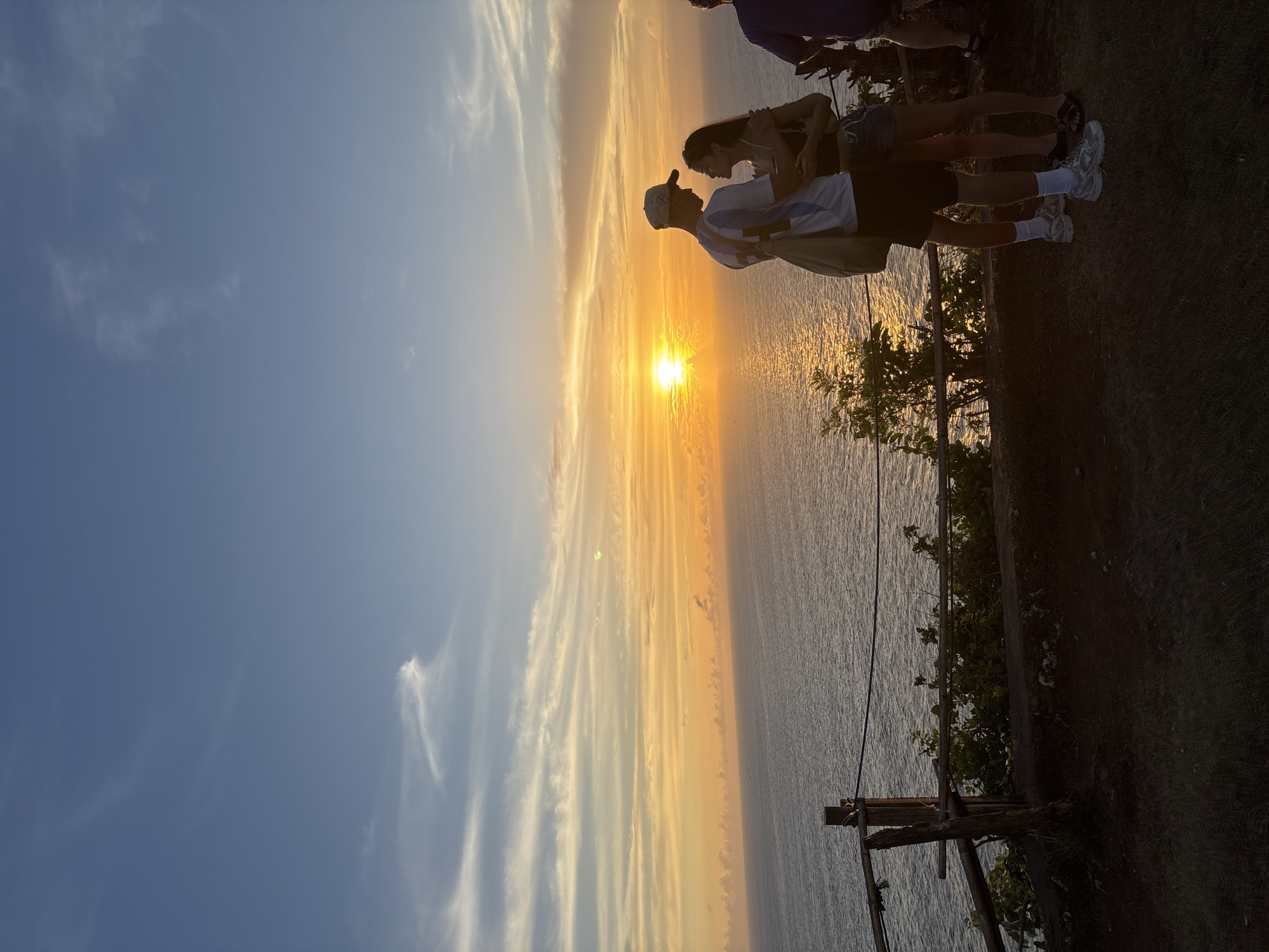 The image depicts a sunset over the ocean, likely in Kuta Selatan, Indonesia, based on the presence of tropical vegetation. A group of people is standing at the edge of a cliff, observing the sunset. A man wearing a cap and a white shirt is standing very close to a woman with long, dark hair. The sun reflects in the ocean with some clouds in the sky. A wooden fence runs along the edge of the cliff.
