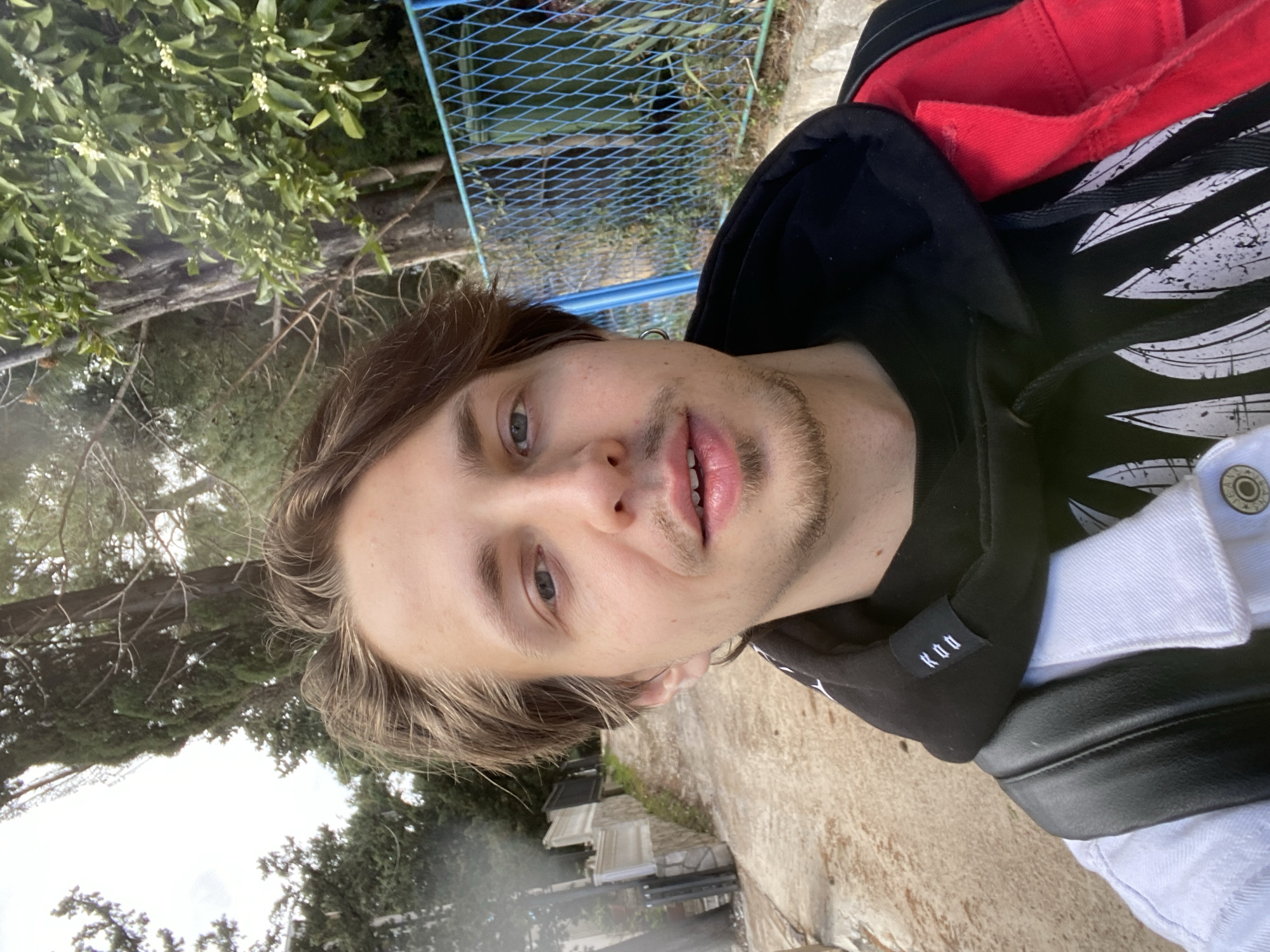 A medium shot of a young adult male, presumably in his late teens to early twenties. He is looking at the camera with a neutral expression. He has light-colored skin, light brown hair, and blue eyes. He has light facial hair. He is wearing a black hoodie with white graphic designs, a red layer underneath, and a white denim jacket. The setting appears to be outdoors, possibly in a park or garden, with greenery and trees visible in the background. There's also a blue metal fence. The ground looks like a paved path or street. The picture was taken during daylight. Based on the location details, the man is likely in Šušanj, Montenegro.