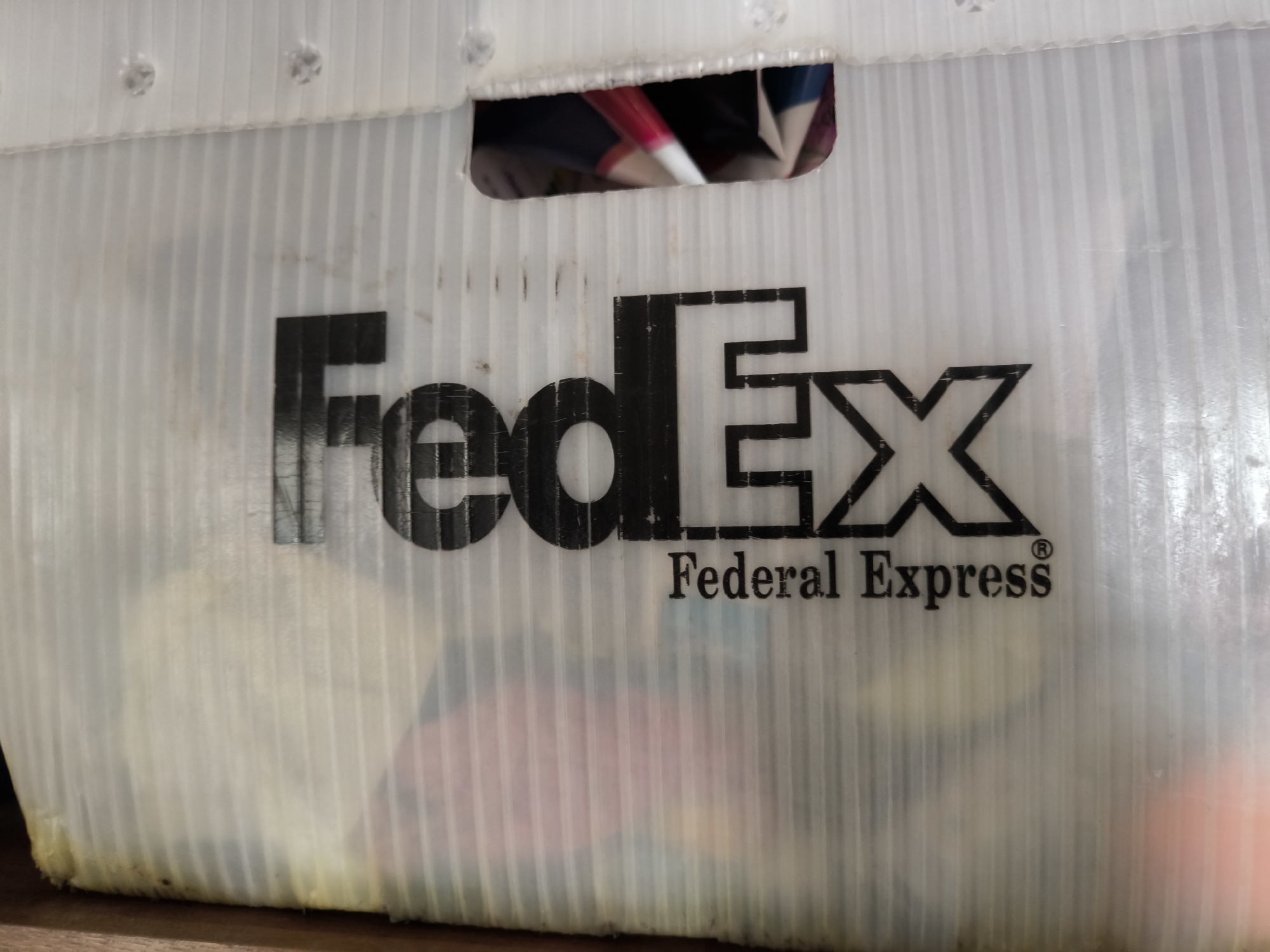 The image displays a close-up, angled view of a white, ribbed plastic bin with the "FedEx Federal Express" logo printed on its side. The logo is in black and consists of the letters "FedEx" in a distinctive font, followed by "Federal Express" in a smaller, serif font, and a registered trademark symbol. The bin appears to be translucent, with various items, some of which are brightly colored fabrics, visible through it. The bin is resting on a wooden surface. There is a rectangular cutout at the top of the bin, through which more of the contents are visible. The overall condition of the bin suggests it is used, with some smudges and discoloration. The scene does not contain any elements that would suggest a specific location in the city of Gal’chino, Russia.