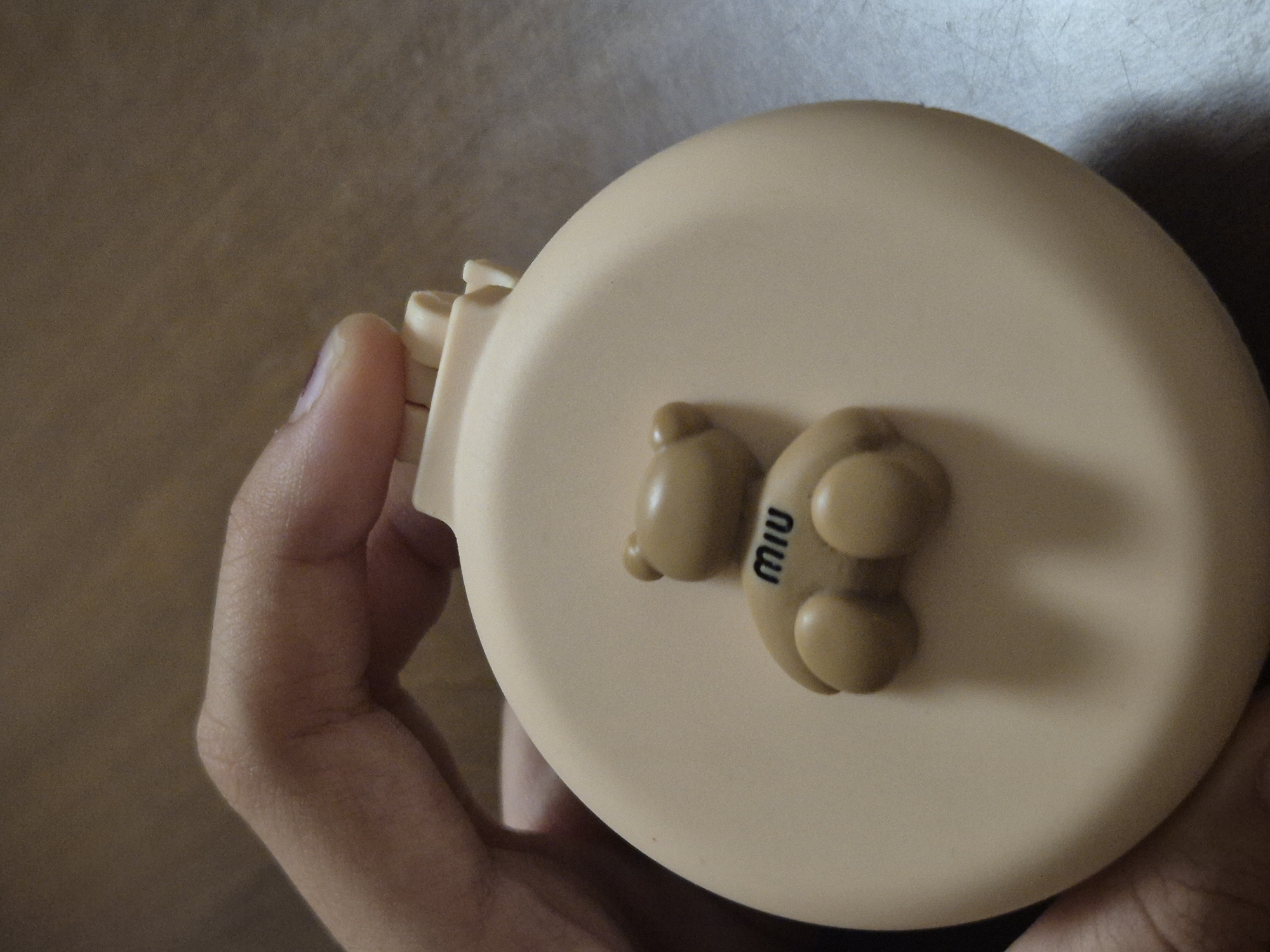 A hand is holding a round, light brown compact mirror. A small, light brown teddy bear figure is attached to the surface of the mirror, with the word "miu" inscribed on its chest in black. The background appears to be a wooden surface. The image is taken from a close-up perspective, focusing on the mirror and the hand. There is no information about the city of Kokshetau, Kazakhstan, in the provided image.