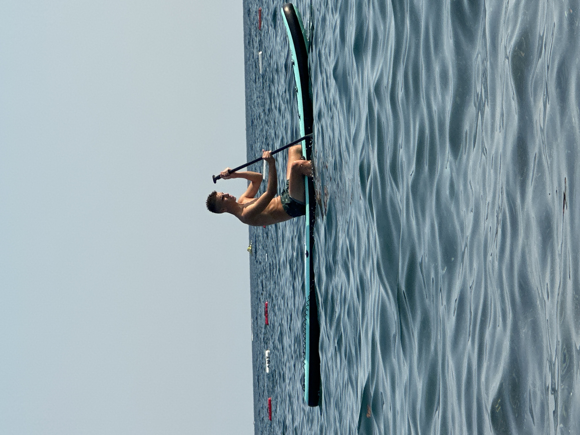 A young man with dark hair is sitting on a paddleboard in the water. He is wearing dark patterned swim trunks and is shirtless. He is holding a paddle in his hands, with the blade submerged in the water. The paddleboard is blue and black. The water is choppy and blue. In the background, there are several buoys floating on the water, and the sky is clear and light blue. The scene appears to be taking place on a sunny day in the sea near Boreti, Montenegro.