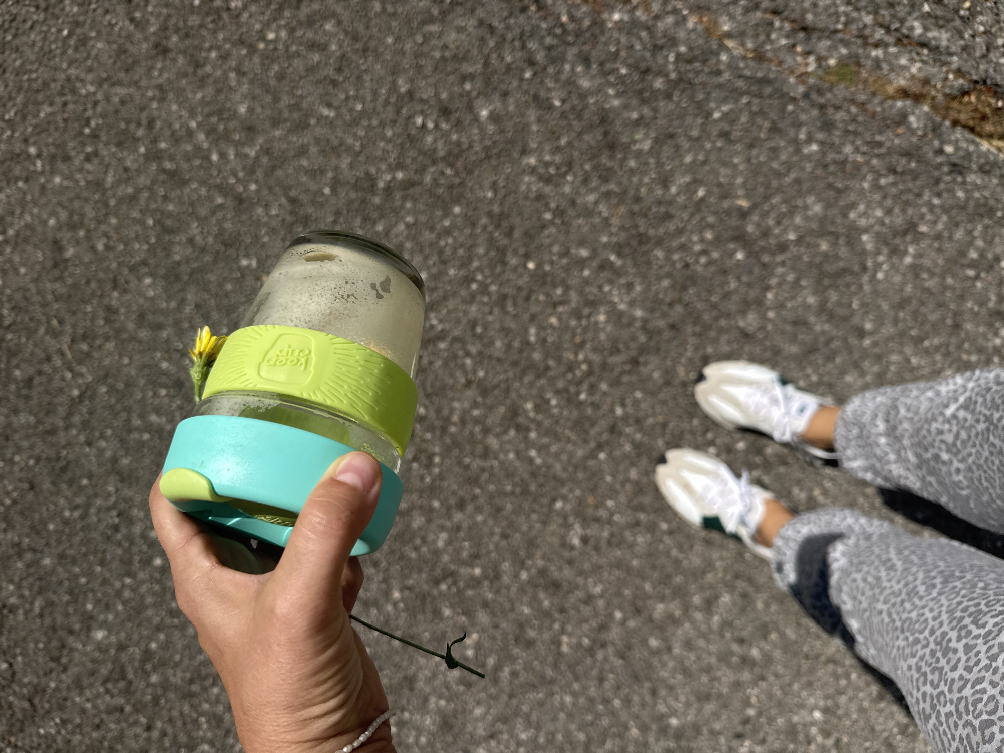 A person stands on a paved surface, likely in Kaluđerac, Montenegro, holding a clear glass jar with green and blue silicone sleeves. The jar appears to contain a liquid and has a yellow flower attached. The person's hand is visible, wearing a white beaded bracelet. The person is also wearing spotted grey pants and white sneakers with green accents. The paved surface is grey and speckled.