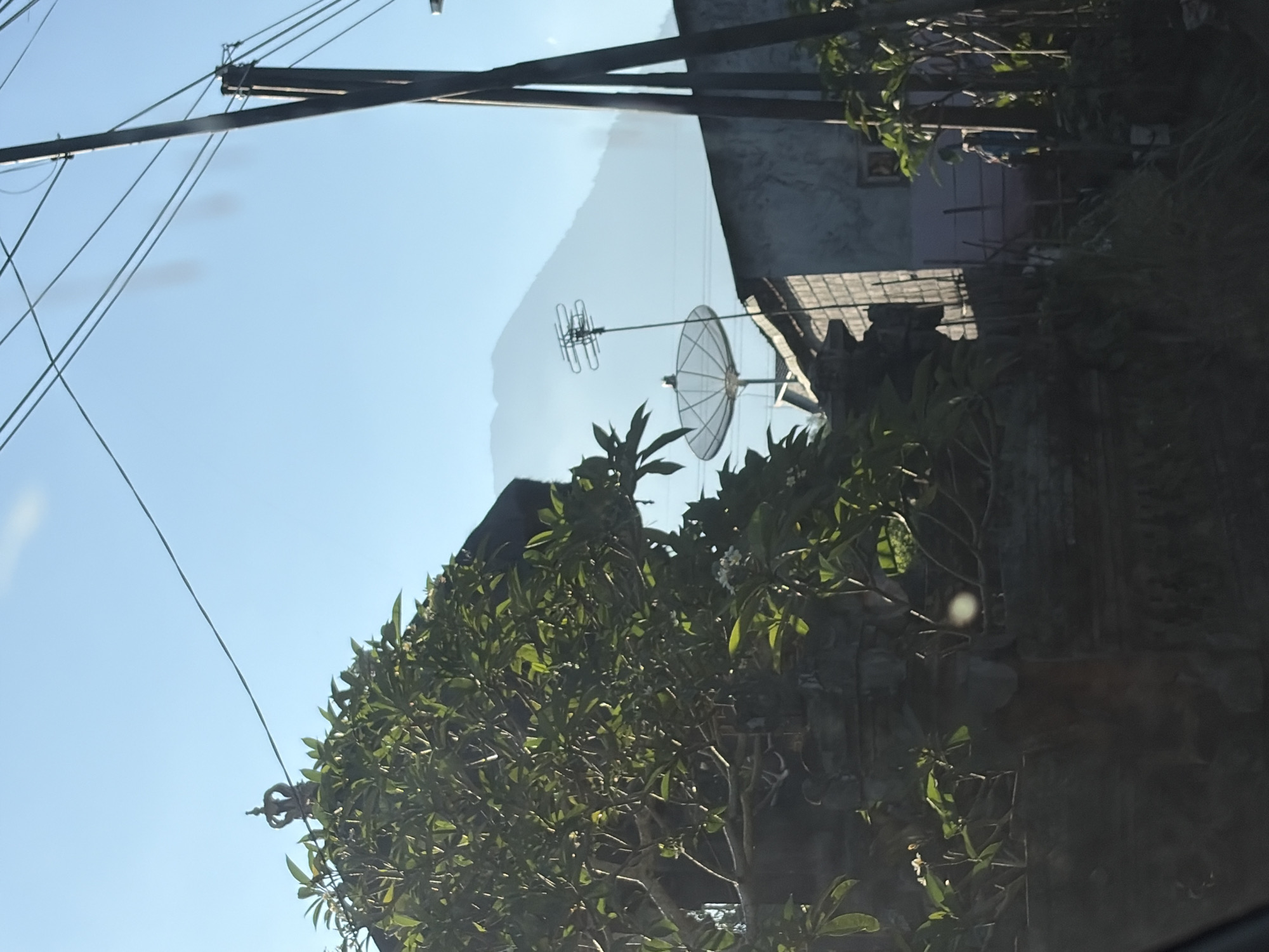 The image captures an outdoor scene, likely in Karangasem, Indonesia, under a clear blue sky. In the foreground, lush green foliage partially obscures a stone structure topped with what appears to be a decorative crown. To the right, a utility pole with multiple wires stretches upwards, with another pole visible further back. Beyond the foliage and structures, a satellite dish and an antenna are mounted, pointing towards a hazy mountain range in the distance. A building constructed with cinder blocks and a tiled roof is visible to the right of the satellite dish. The overall impression is of a residential or semi-rural environment with a backdrop of natural landscape.