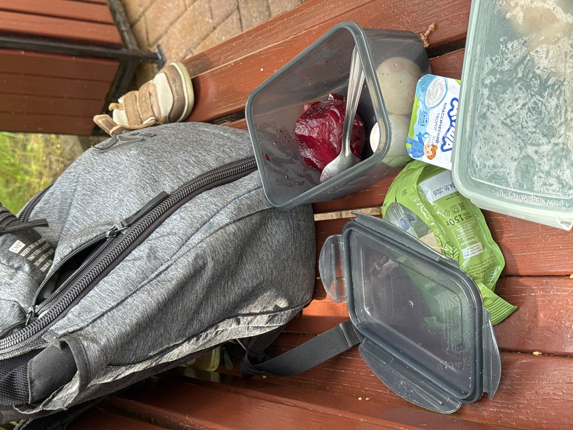 The image shows a gray backpack and various food containers resting on a red-brown wooden bench. There is a small tan and white children's shoe near the backpack. The food containers include a clear plastic container with condensation inside, a gray container holding cooked beets and a spoon, a white container with blue lettering, and a green snack package. The label on the snack package includes the text "150 г", a production and packaging date of 04.05.2026, and contact information for quality control. The white container has text in Cyrillic characters that include the words "КЛАССИЧЕСкий ТВОРОГ" and "Ca+K2". The location is Estosadok, Russia.